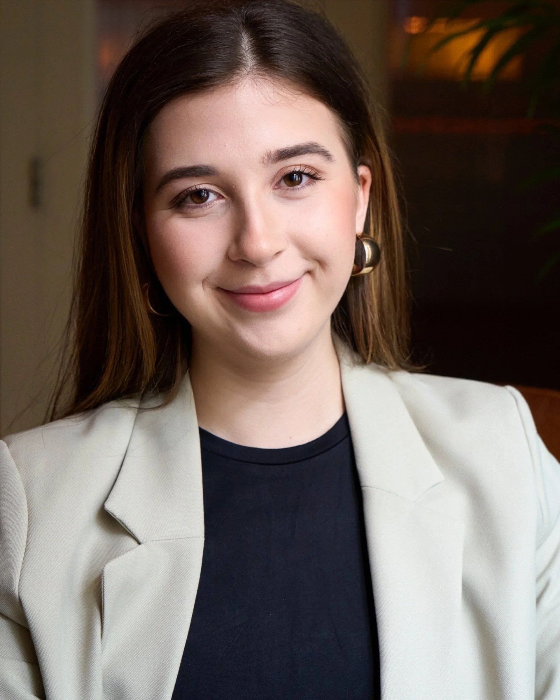 A young woman with brown hair and wearing a black top and beige blazer smiling at the camera.