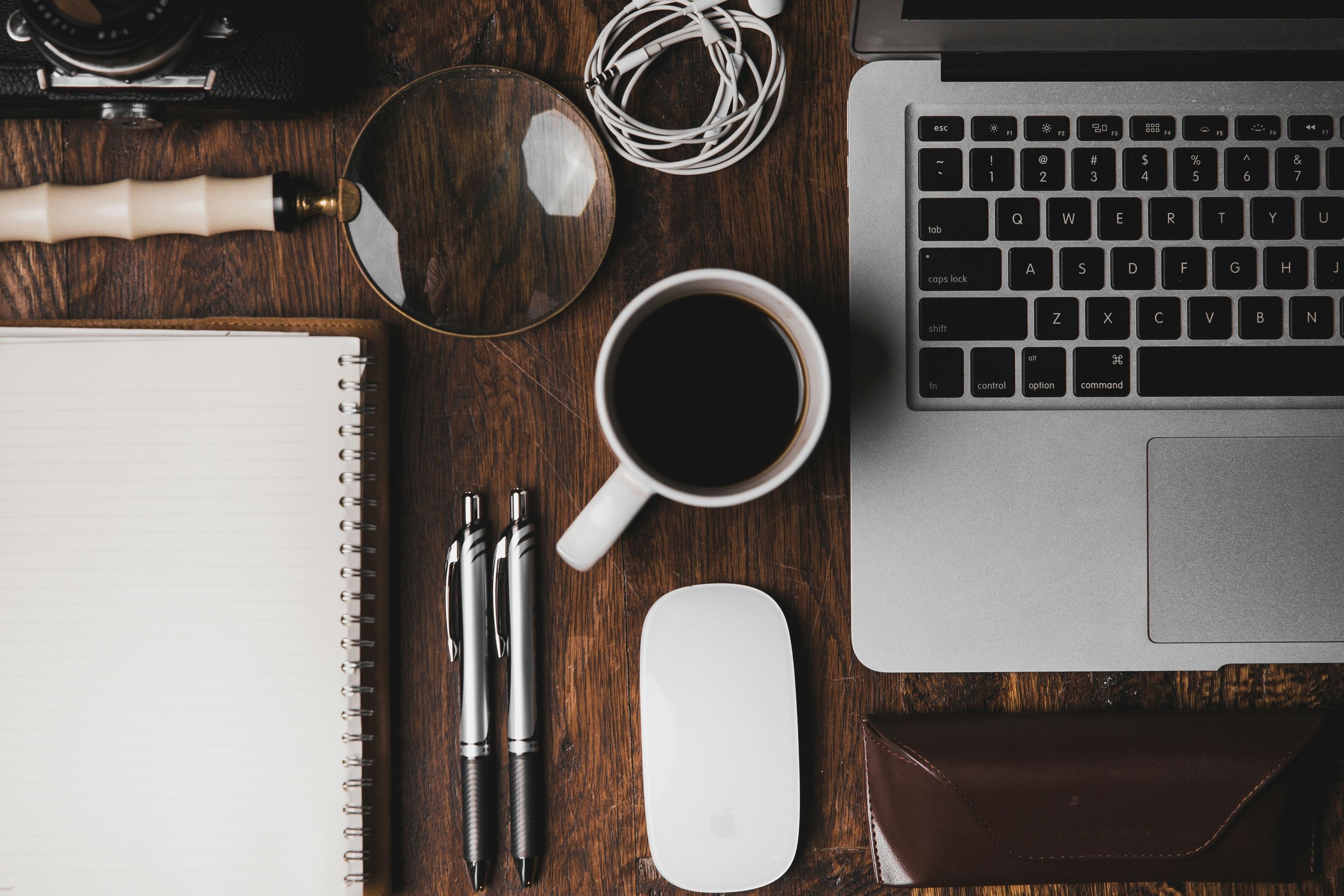 A wooden desk with a camera, tangled white earbuds, a laptop, a notepad with pens, a cup of black coffee, a circular mirror, and a few black markers.
