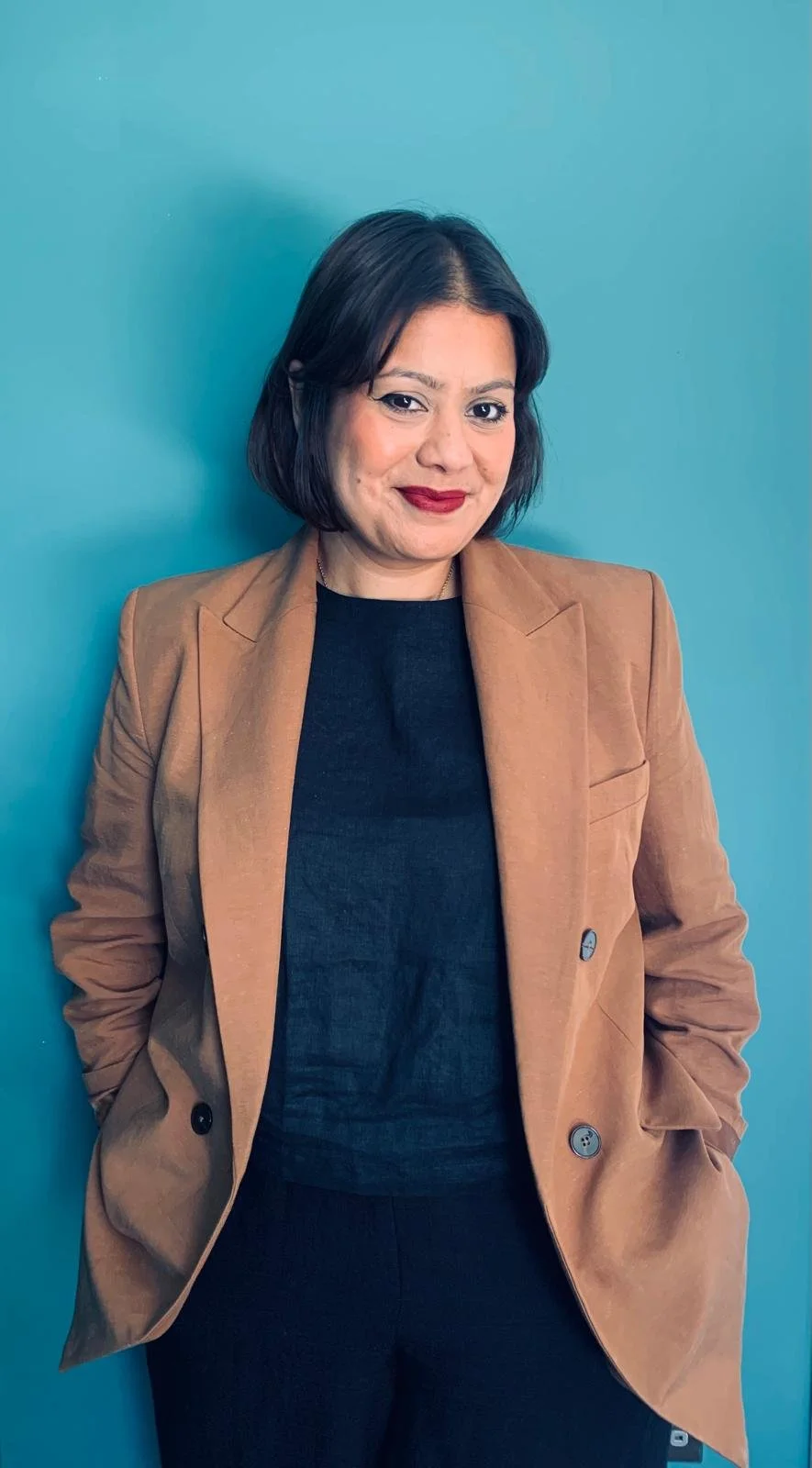 A woman with shoulder-length black hair, wearing a tan blazer over a black top, standing against a blue background.