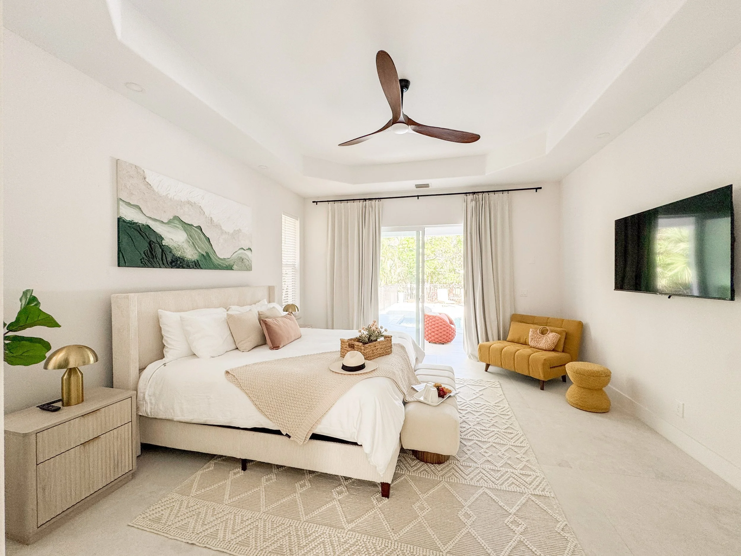 Bright and airy bedroom with white walls and bedding, a yellow upholstered chair, a bed with beige and pink pillows, a wooden nightstand with gold lamps, a large abstract green and white painting, a sliding glass door with beige curtains, and a mount