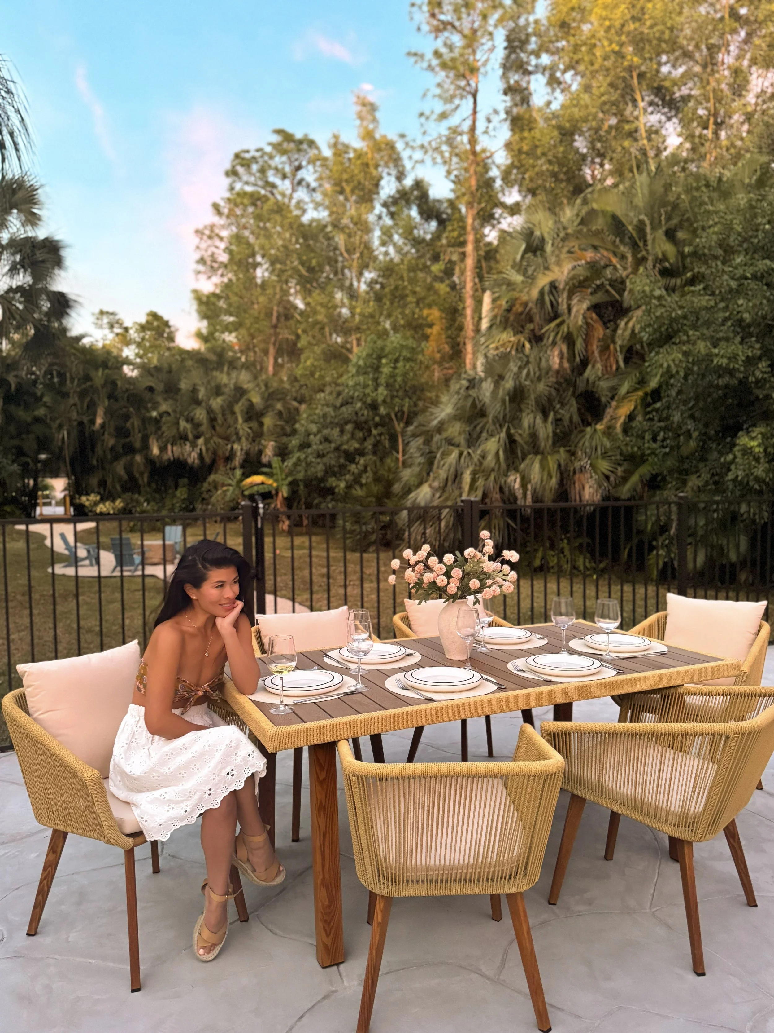 A woman in a white lace skirt and beige high heels is sitting at an outdoor dining table with tableware and a vase of pink flowers, surrounded by wicker chairs and greenery, with trees and a blue sky in the background.