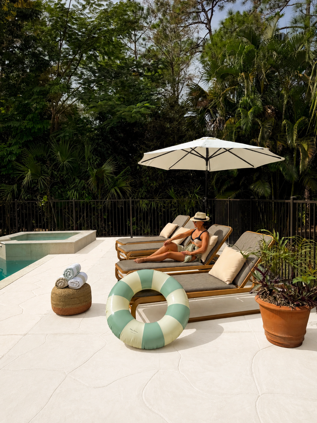 A woman relaxing on a sun lounger by a backyard pool, shaded by a large white umbrella, with tropical green trees and plants in the background, surrounded by poolside accessories including rolled towels, a life preserver, and a potted plant.