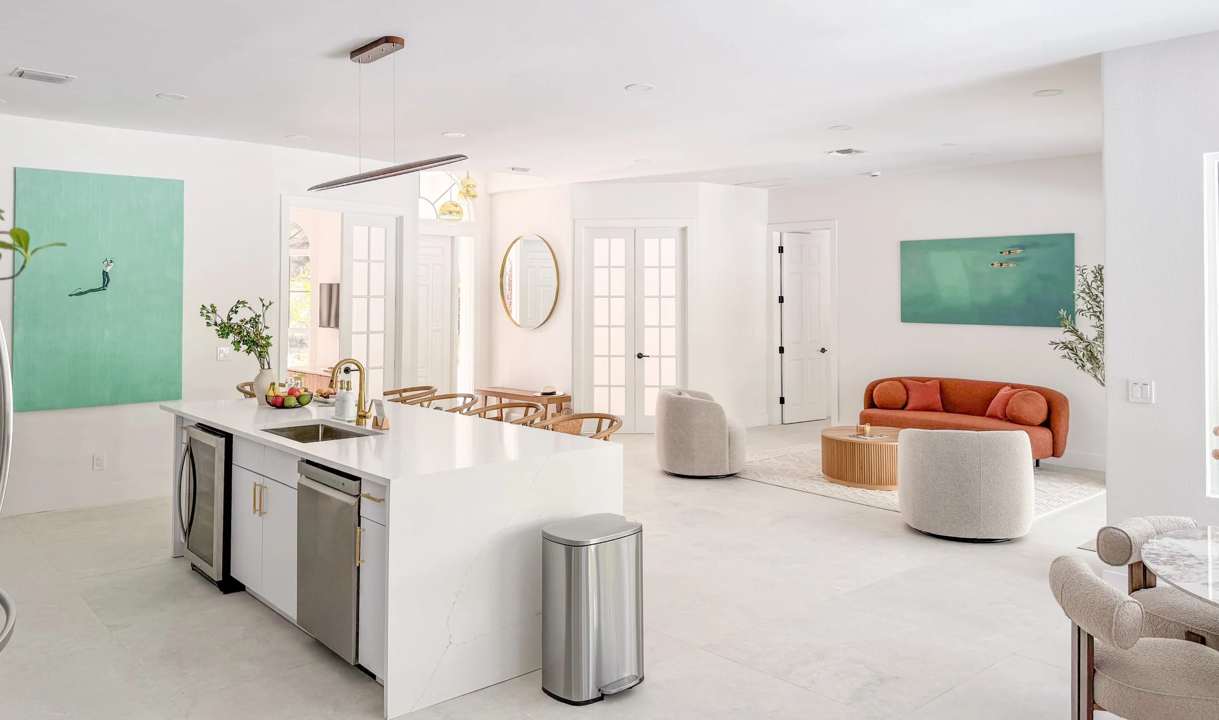 Bright, open-concept living room and kitchen with white walls and modern furniture, artwork, greenery, and natural light.