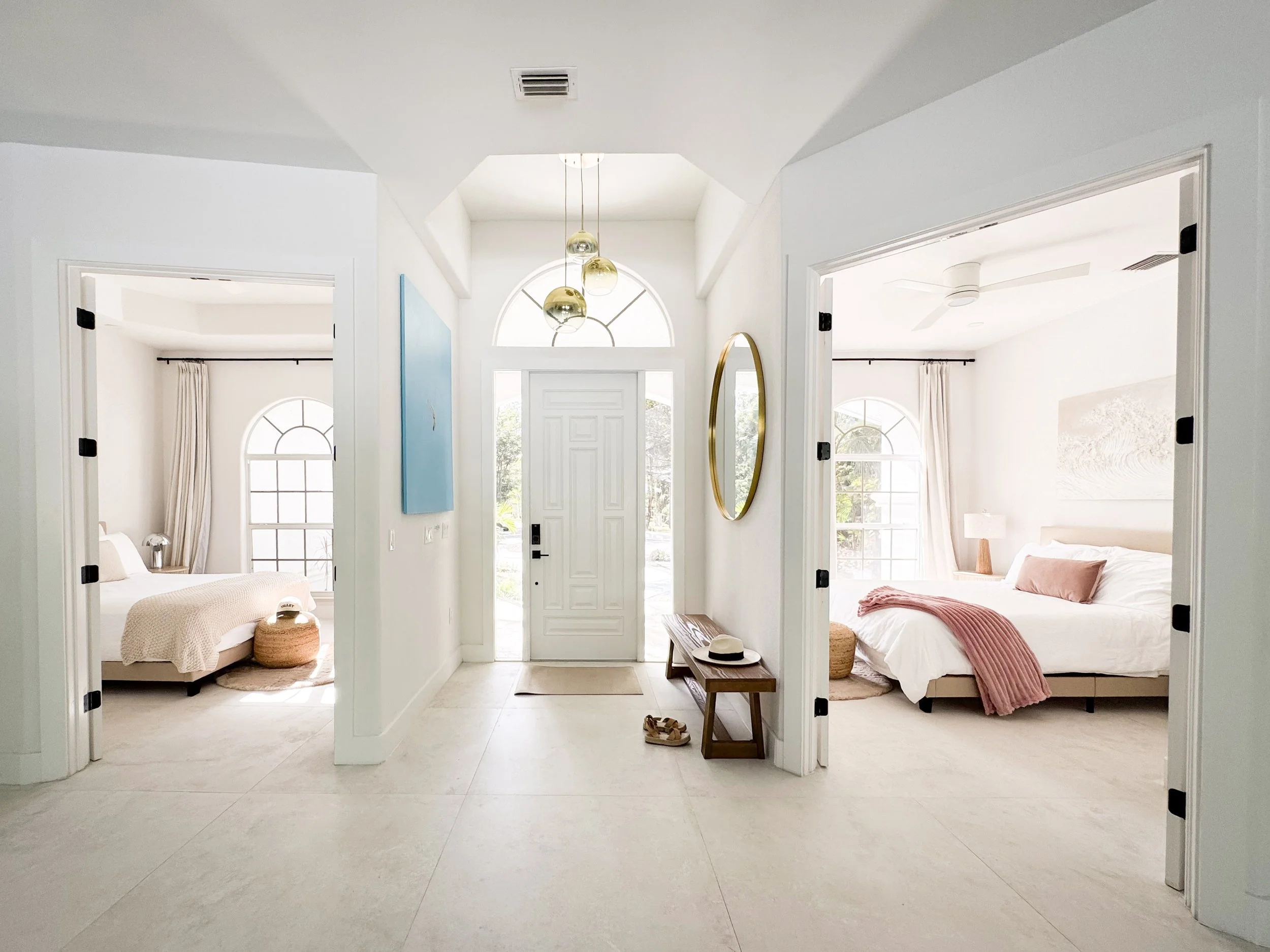 Interior view of a bright, modern home with two bedrooms, a front door, and large windows, featuring white walls, light-colored flooring, and minimal decor.