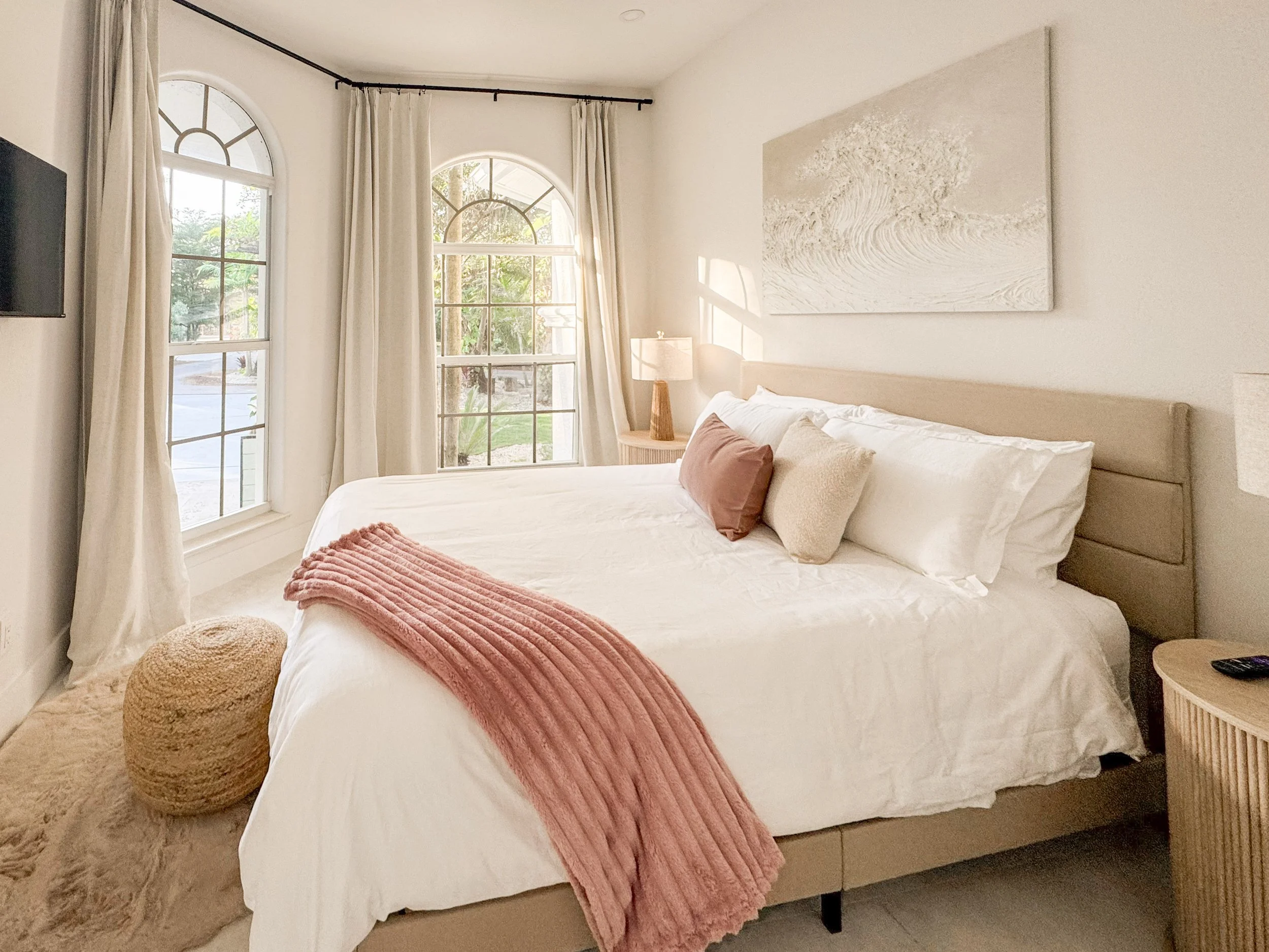 A cozy bedroom with a bed against a wall, dressed in white linens and decorated with beige, cream, and dusty pink pillows. A pink throw blanket is draped along the foot of the bed. To the left of the bed is a textured rounded beige pouf. Behind the b