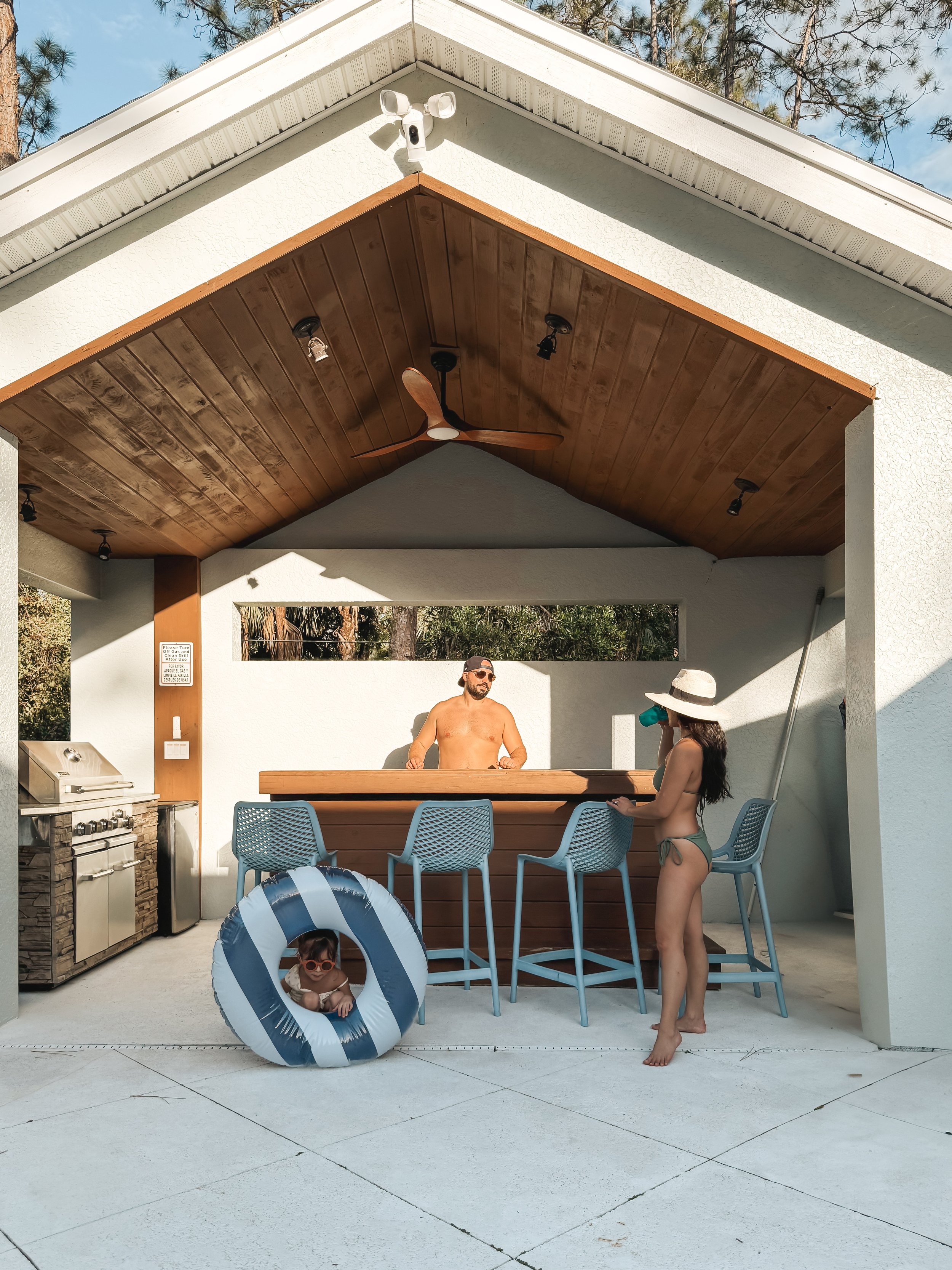 A man with a beard and sunglasses stands shirtless behind an outdoor bar counter with a woman in a swimsuit and sunhat in front. A young child with sunglasses is inside a small inflatable pool on the ground in front of the bar, and another child with
