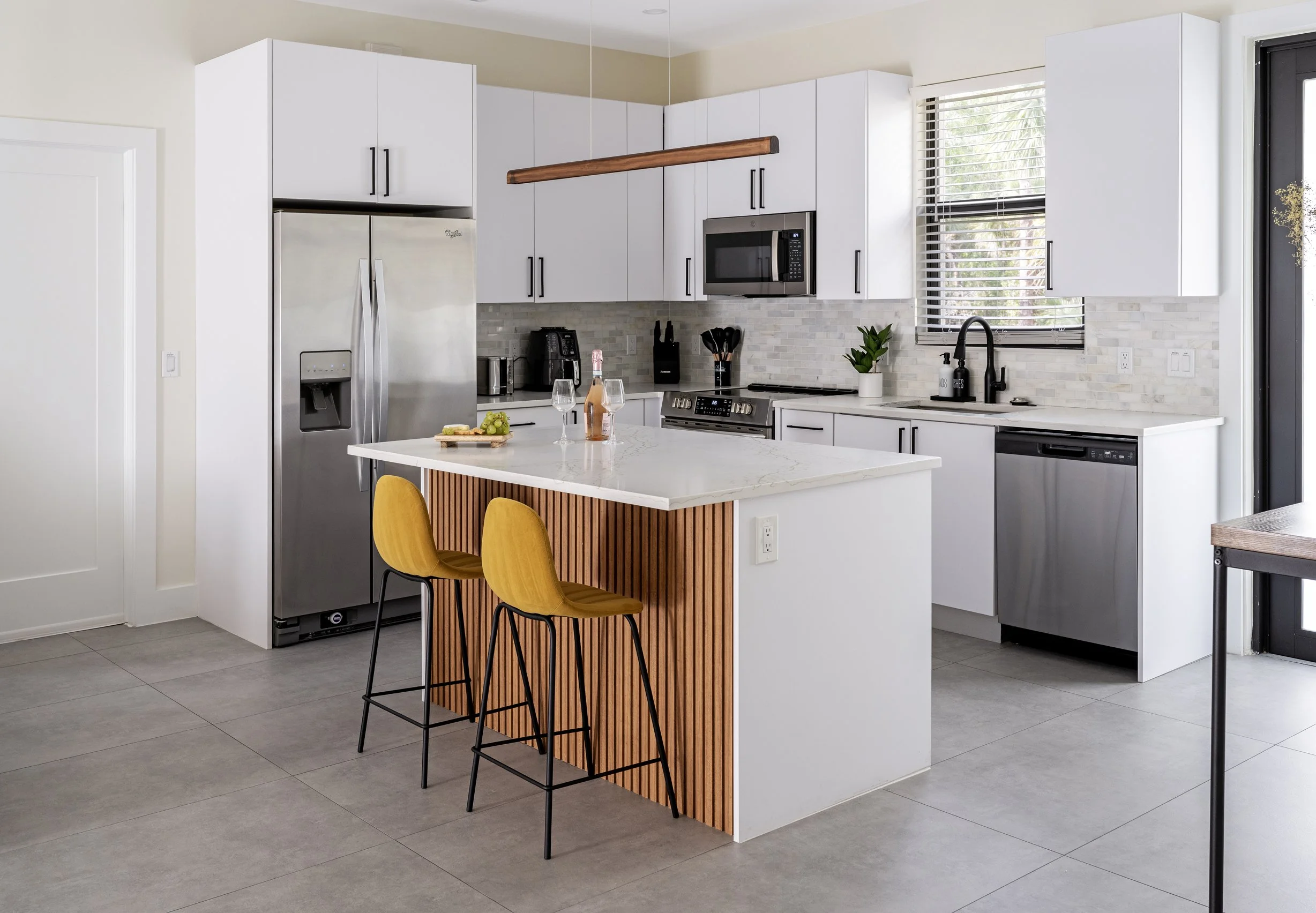 A modern kitchen with white cabinets, a stainless steel refrigerator, microwave, and oven, a white marble island with yellow bar stools, and gray tiled flooring. There are small appliances, utensils, and a plant on the countertop, with windows allowi