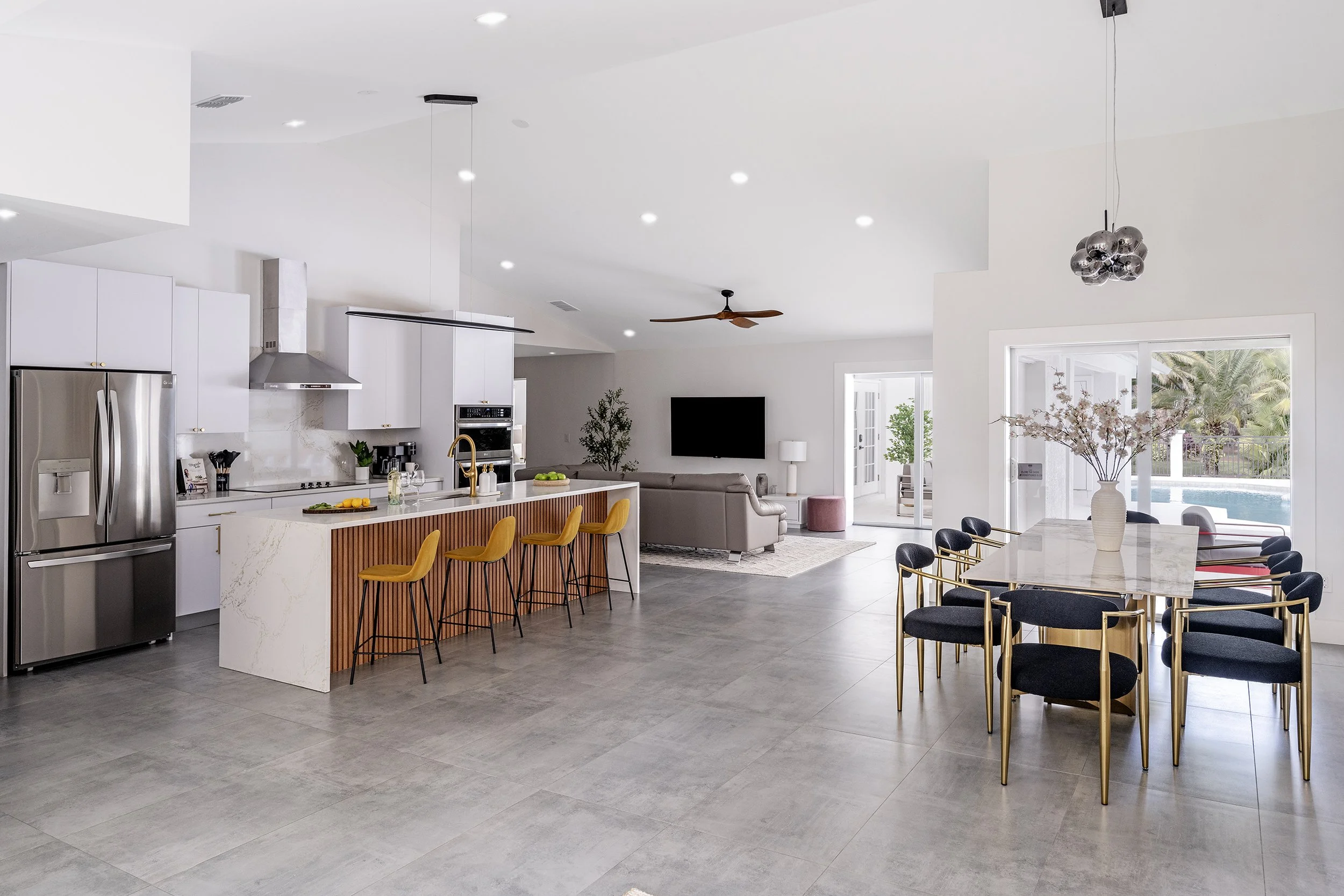 Open-concept living area with a modern kitchen, dining area, and living room. The kitchen features white cabinets, a marble island with yellow stools, stainless steel appliances, and a glass jar with lemon slices. The living room has a light grey sof