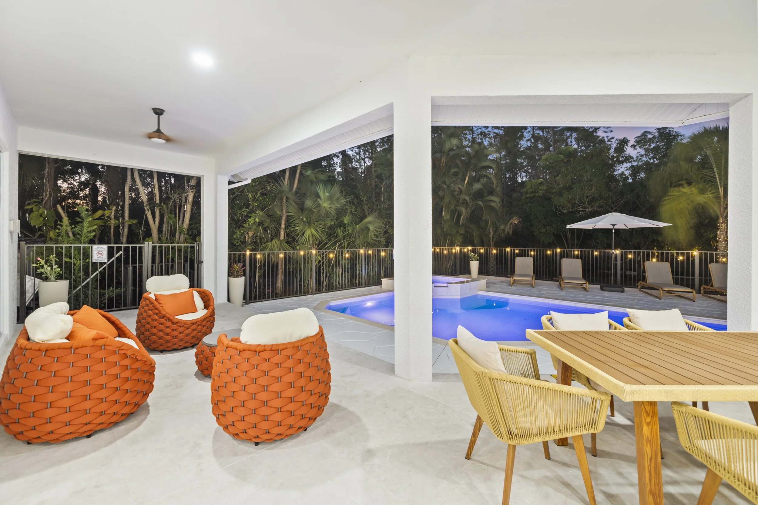 The image shows an outdoor patio with a swimming pool, surrounded by lounge chairs, an umbrella, and lush green trees in the background. There are also some orange and beige cushioned chairs and a table on the patio.