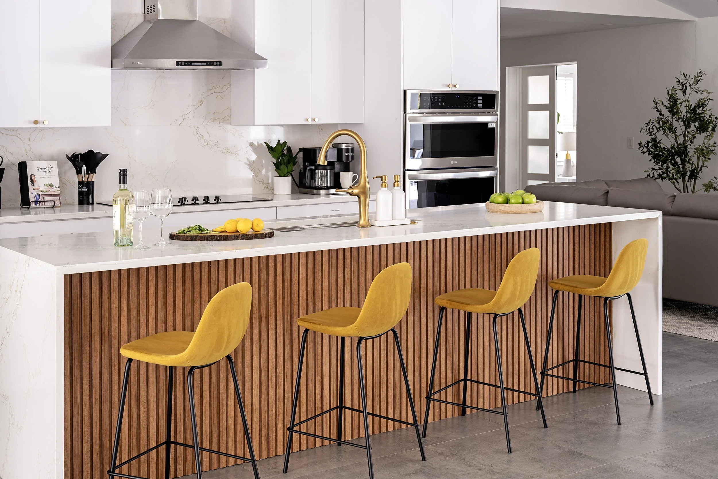 A modern kitchen with yellow barstools at a white marble island, featuring a gold faucet, lemons, and kitchen appliances, with a living room visible through an open doorway.