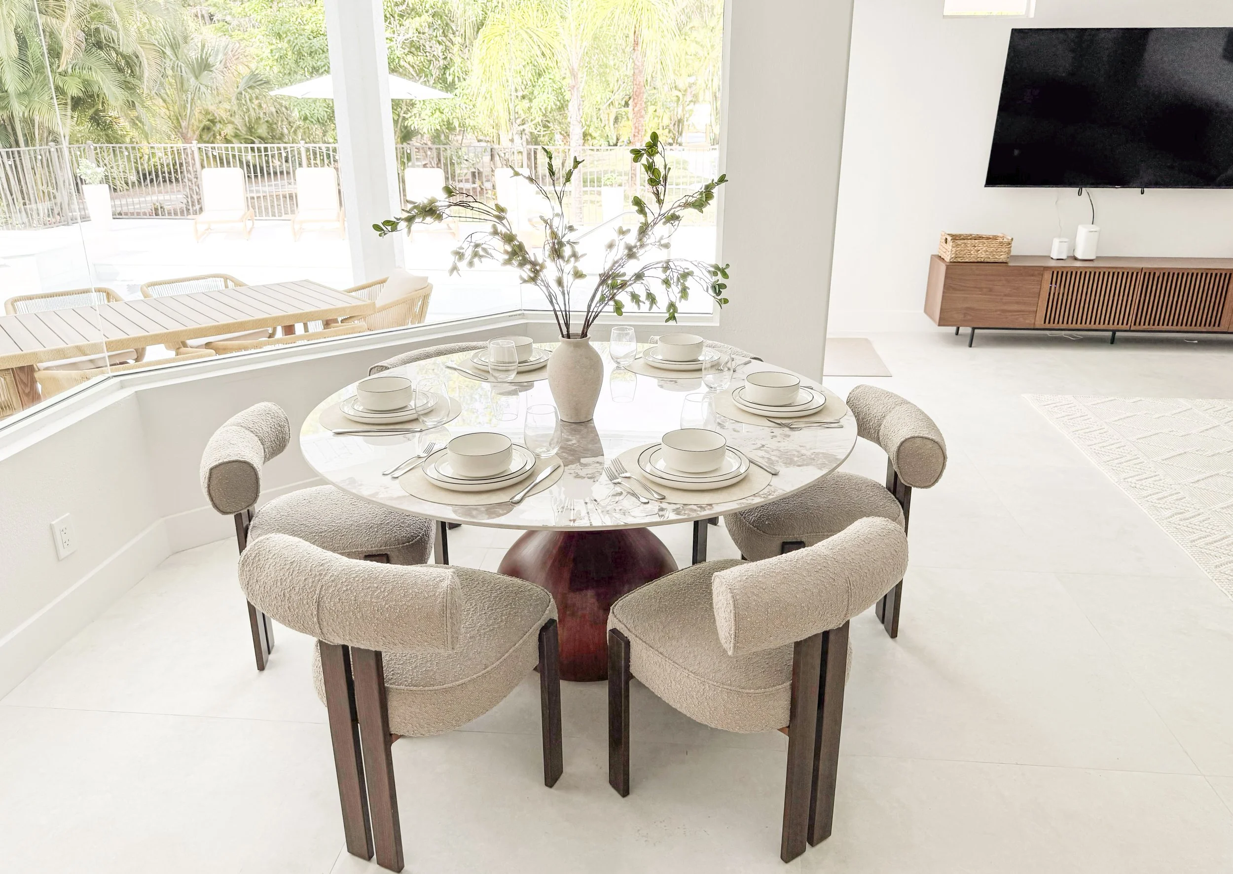 A round dining table set for six with white plates, bowls, and glasses, decorated with a large beige vase containing green branches, surrounded by plush beige chairs, with a large window showing an outdoor patio with lounge chairs and lush greenery i