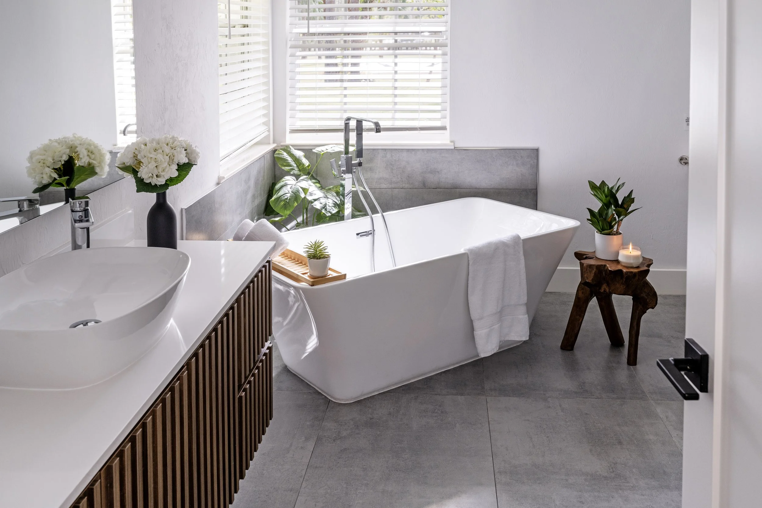 A modern bathroom with a white bathtub, a vanity with white sinks and wooden slats, and decorative elements including white flowers, green plants, and candles. The room has large windows with white blinds and gray tiled floors.