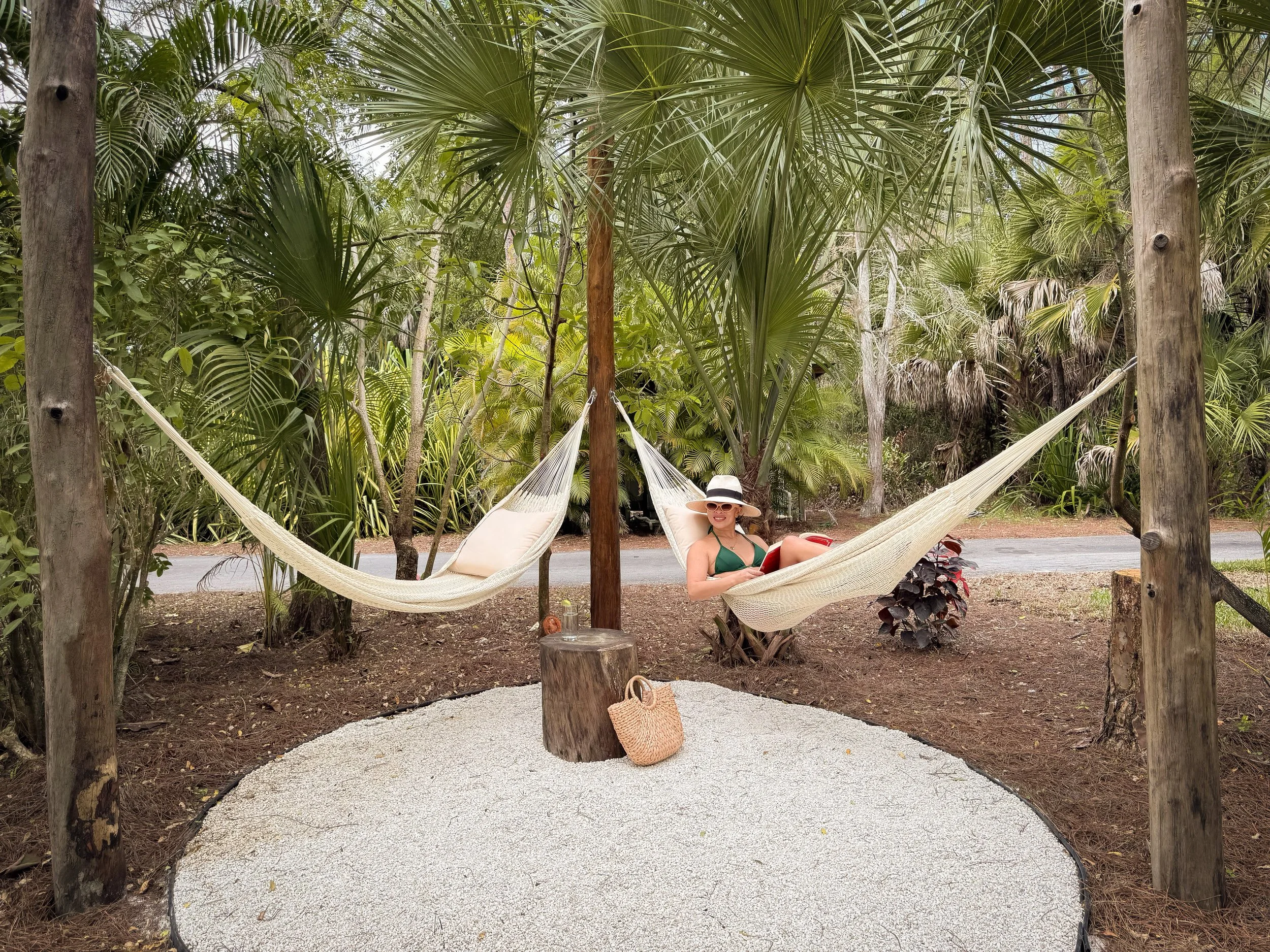 Woman in a green swimsuit and wide-brimmed hat relaxing on a hammock in a lush tropical garden with tall trees and greenery.