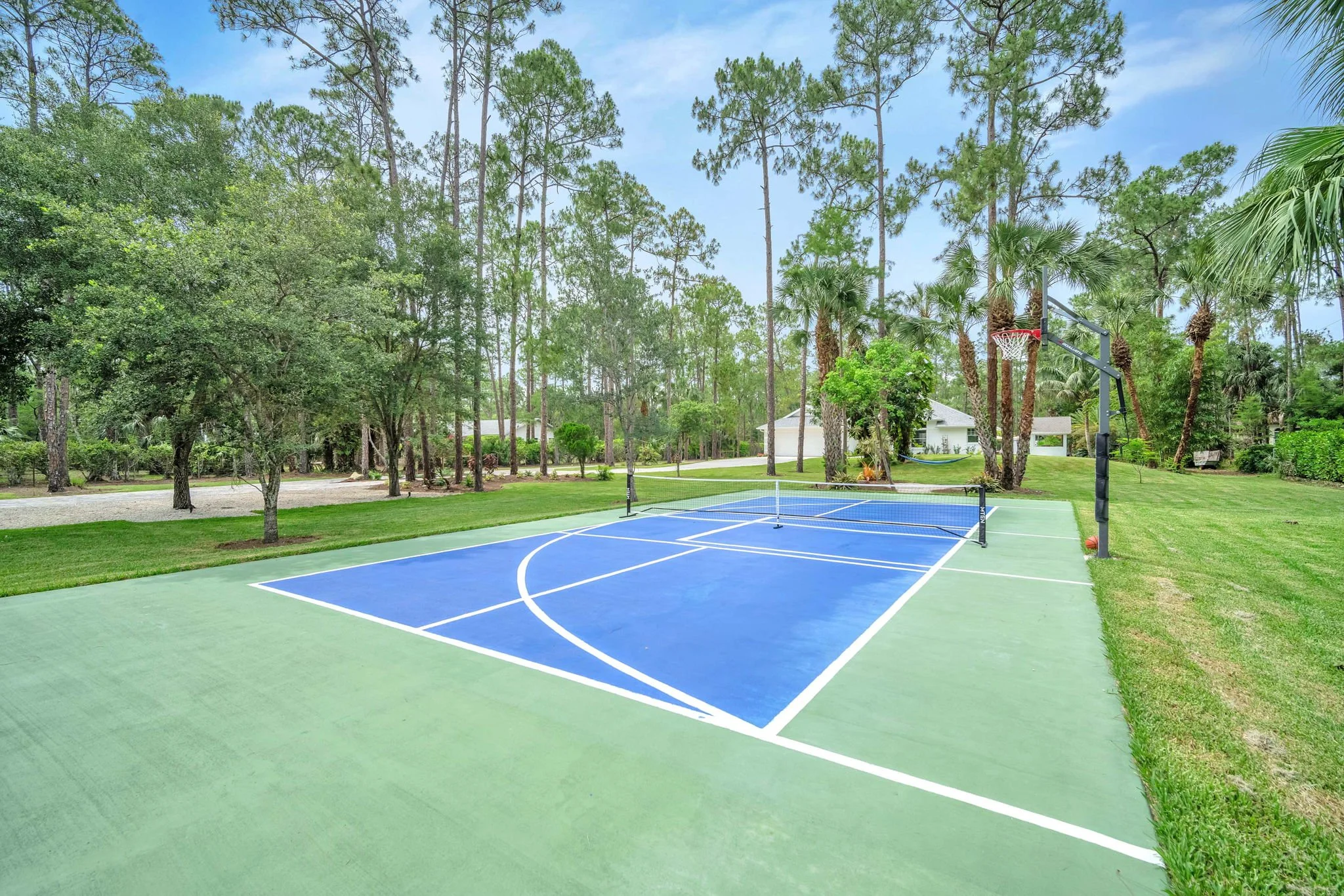 Outdoor basketball court with a blue playing surface, surrounded by lush green trees and a house in the background.