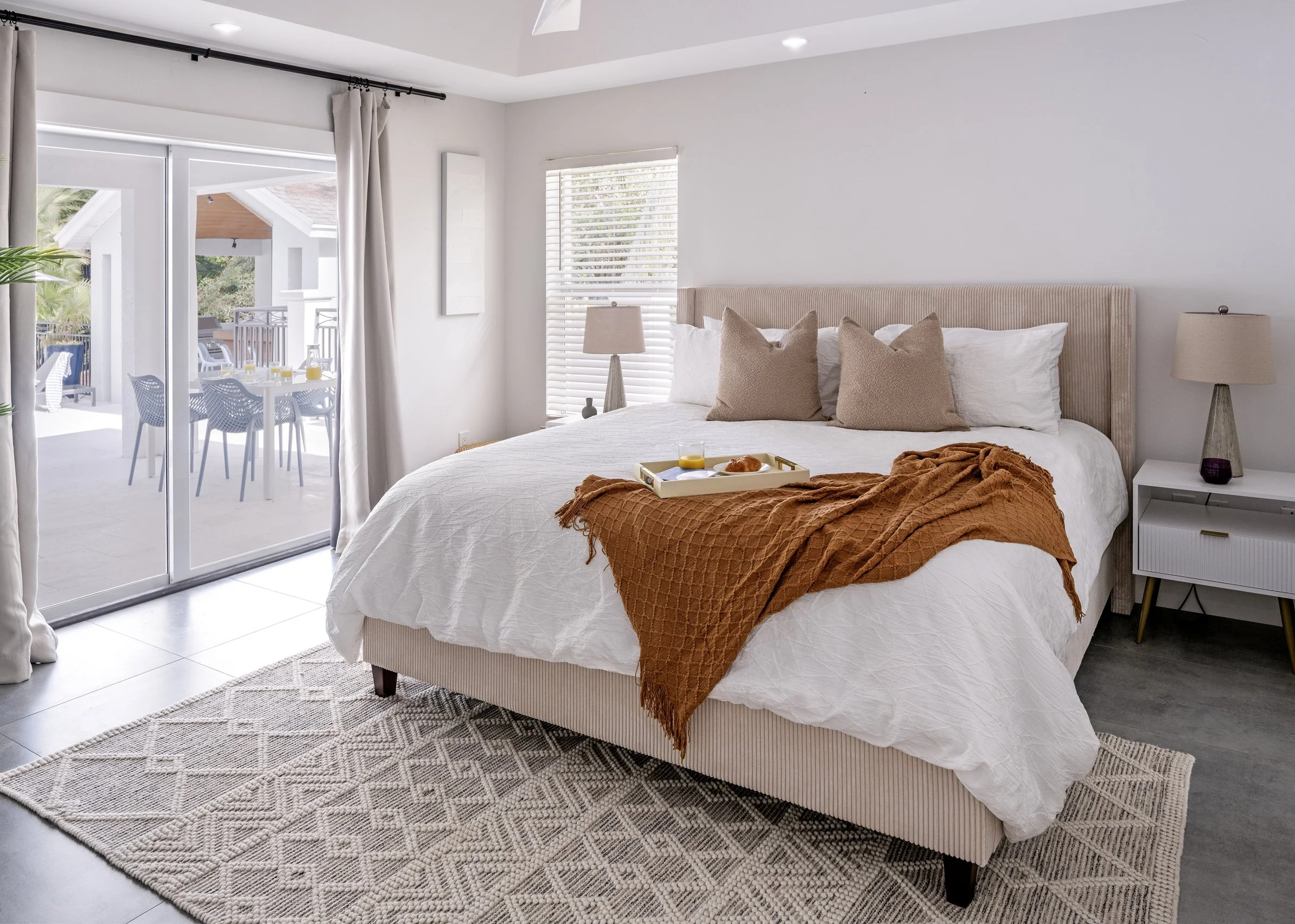 Bedroom with a large bed, beige headboard, white bedding, and pillows. Bed has an orange blanket and breakfast tray with juice and pastries. Nightstand with a beige lamp, window with white blinds, sliding glass door opening to a balcony with outdoor 