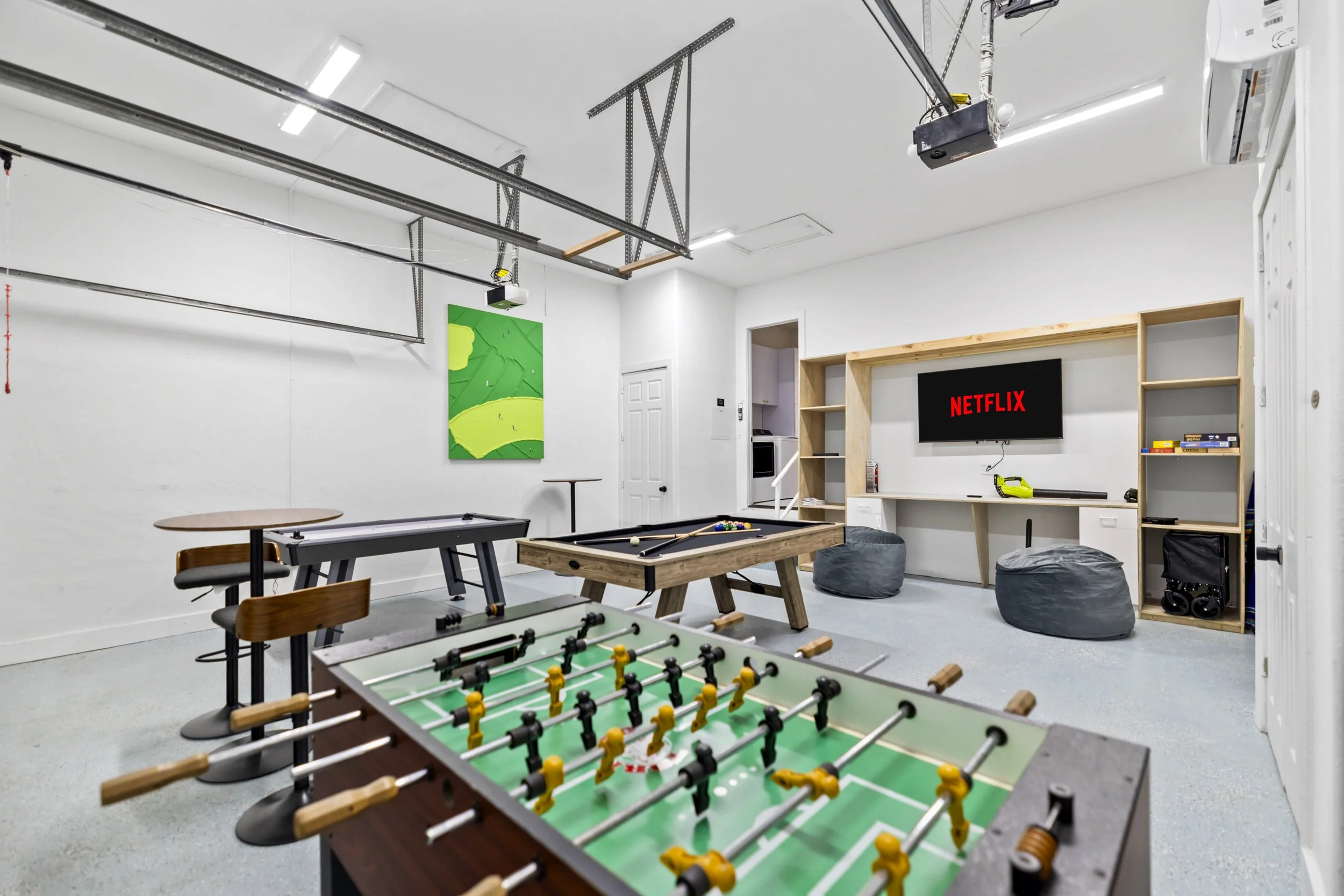 Indoor recreational room with a foosball table in the foreground, a pool table behind it, a wall-mounted TV with the Netflix logo, and a corner bookshelf with books and electronic devices. The room has white walls, ceiling with exposed pipes and equi