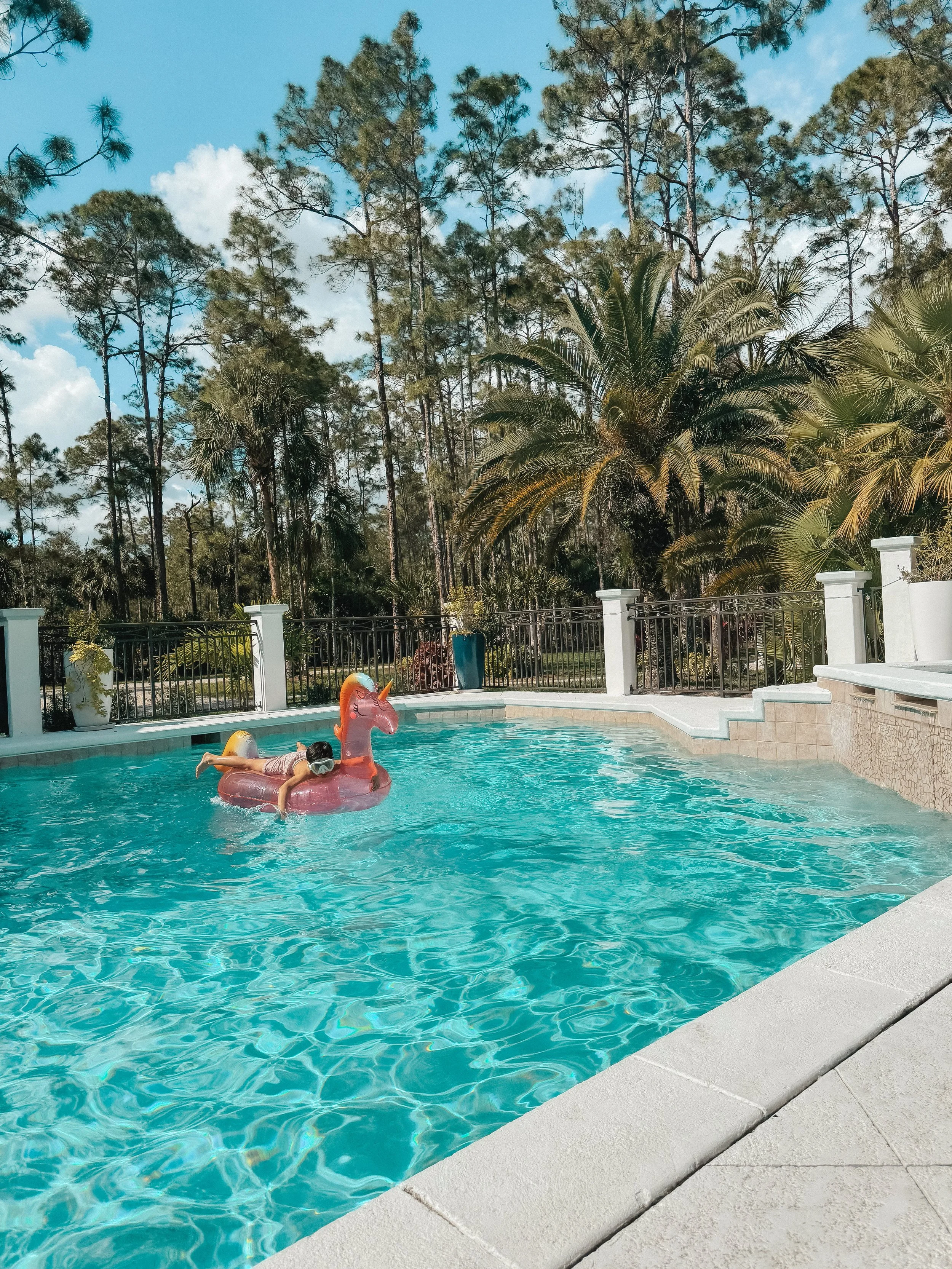 Child floating on a pink inflatable unicorn in a swimming pool, surrounded by a white fence with palm trees and pine trees in the background, under a blue sky with some clouds.