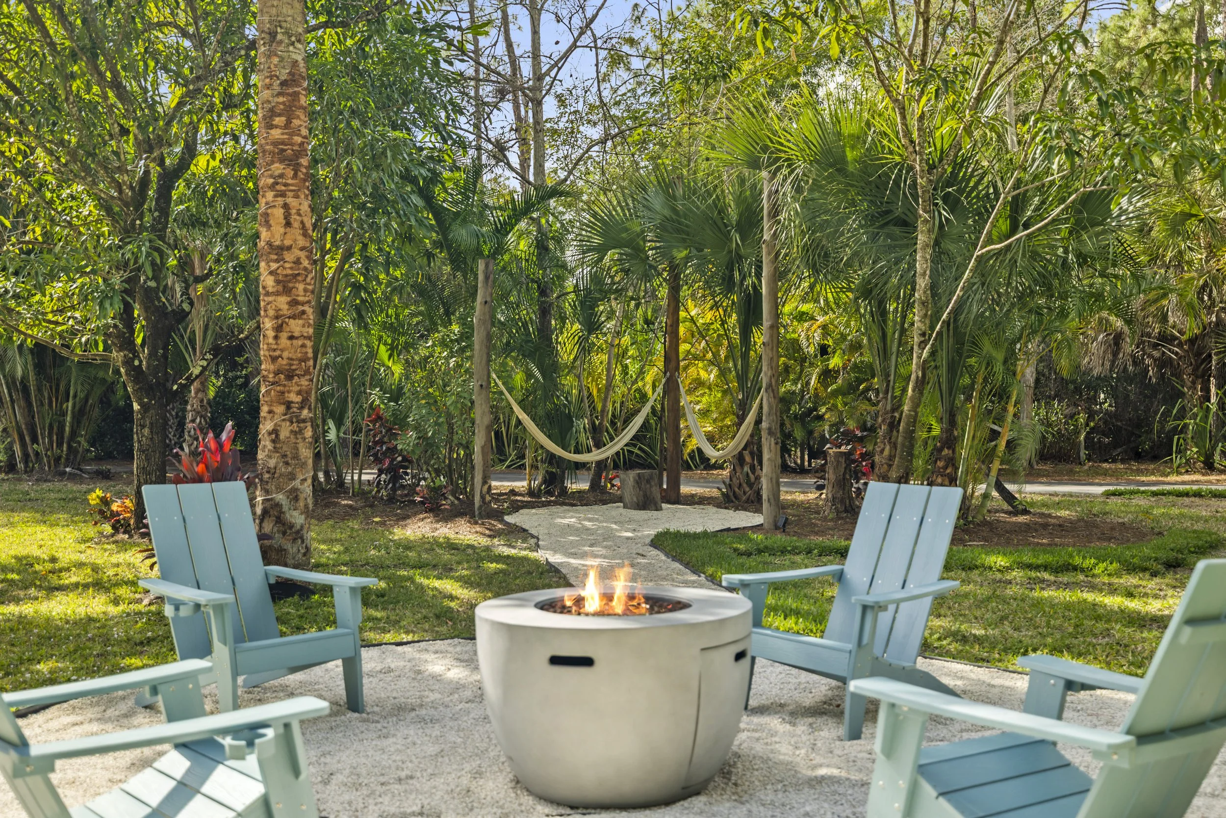 A backyard garden seating area with four light blue chairs arranged around a concrete fire pit with a small flame. There is a fire pit in the foreground and lush green trees and plants in the background, with a pathway and a hammocks among the trees.