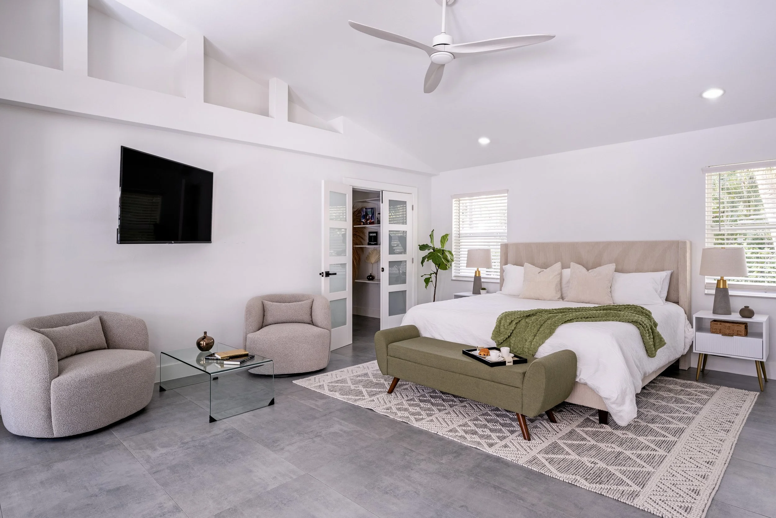A modern, minimalist bedroom with a white bed, beige headboard, green blanket, and wooden legs. There are two nightstands with lamps, a bench at the foot of the bed, a flat-screen TV on the wall, and a glass coffee table with a vase and books. Two be