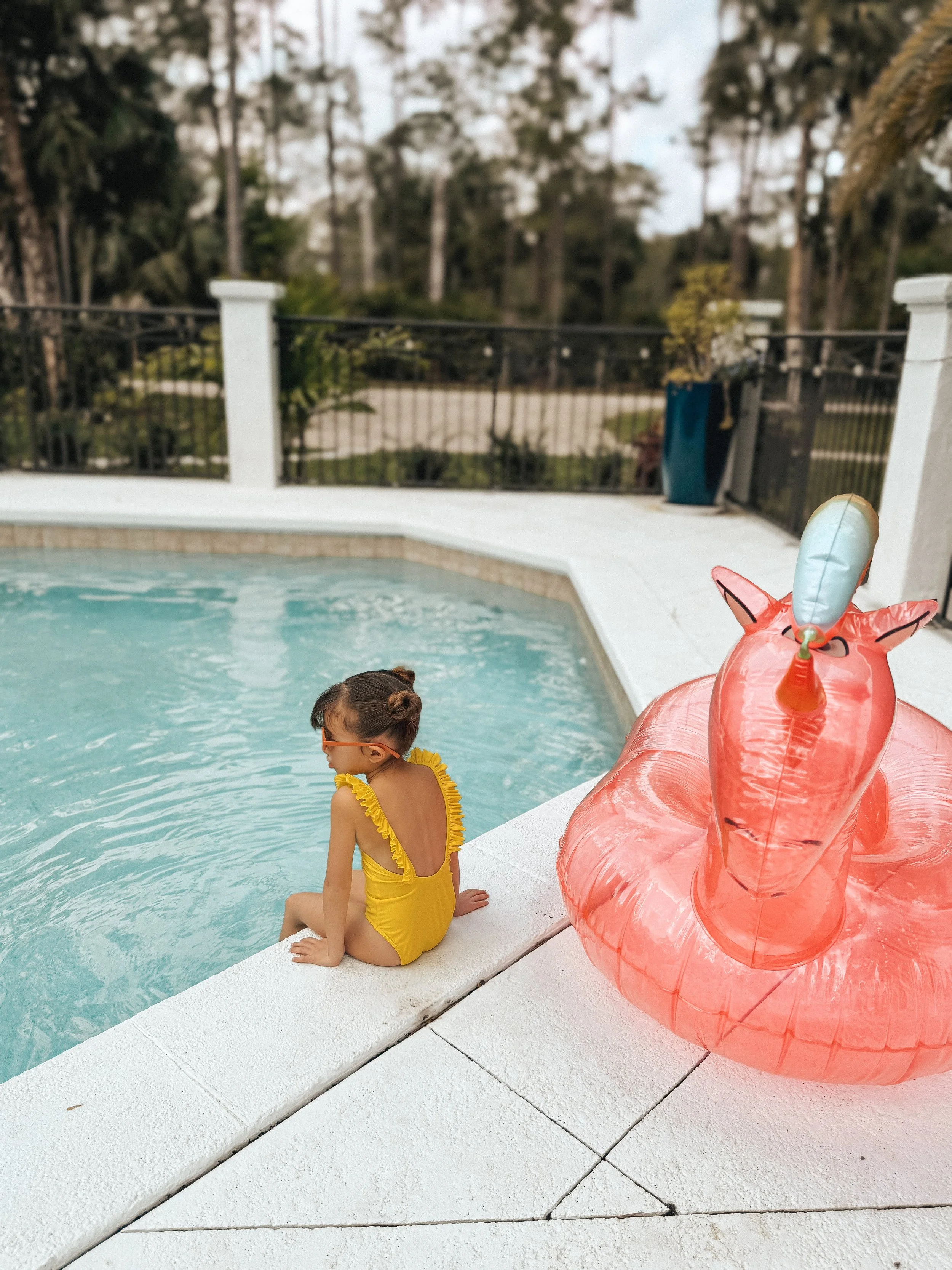 A young girl in a yellow swimsuit sits by the edge of a swimming pool, with her back to the camera, near a large inflatable pink flamingo pool float.