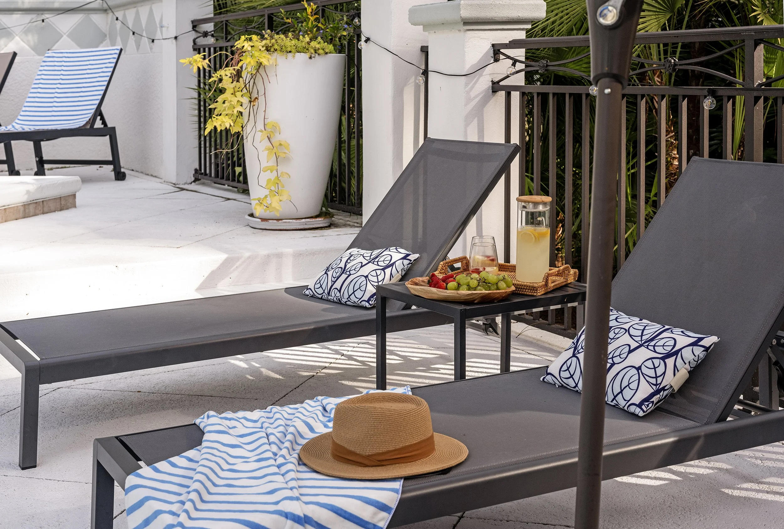 Outdoor patio with black lounge chairs topped with blue and white striped pillows, a round straw hat, a rectangular tray with green grapes and strawberries, a glass of water with lemon slices, a glass jug with lemon water, a large white planter with 
