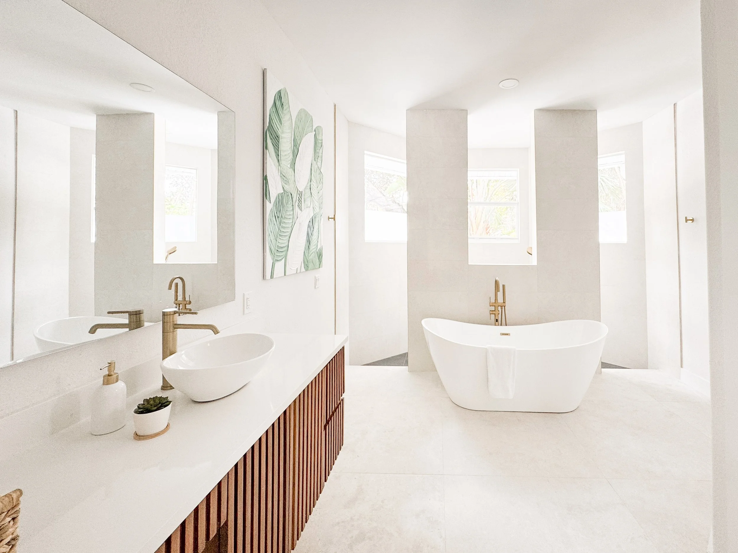 A modern, minimalist bathroom with a white bathtub, double sinks with white vessel basins, gold faucets, a mirror, and a piece of artwork with green leaves on the wall. There are windows letting in natural light.
