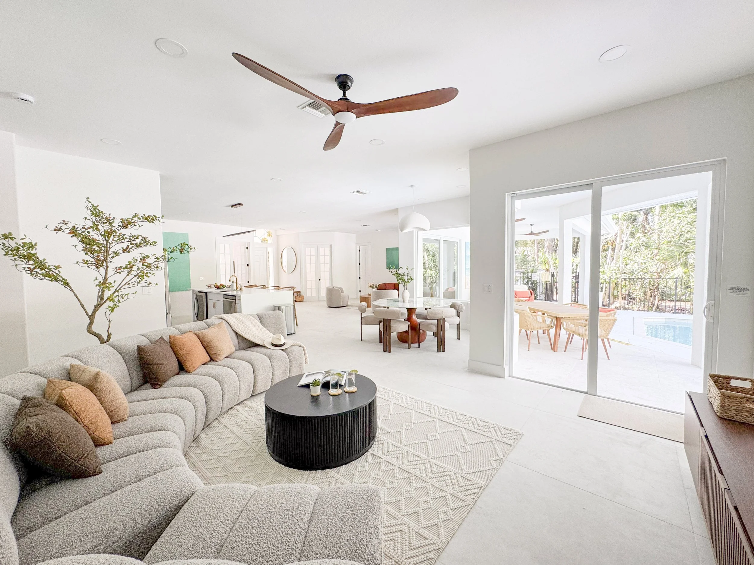 Bright open living room with beige sectional sofa, black round coffee table, and light-colored rug. Dining area with round table and chairs, kitchen with white cabinets, and an outdoor patio with wooden furniture and a pool visible through sliding do