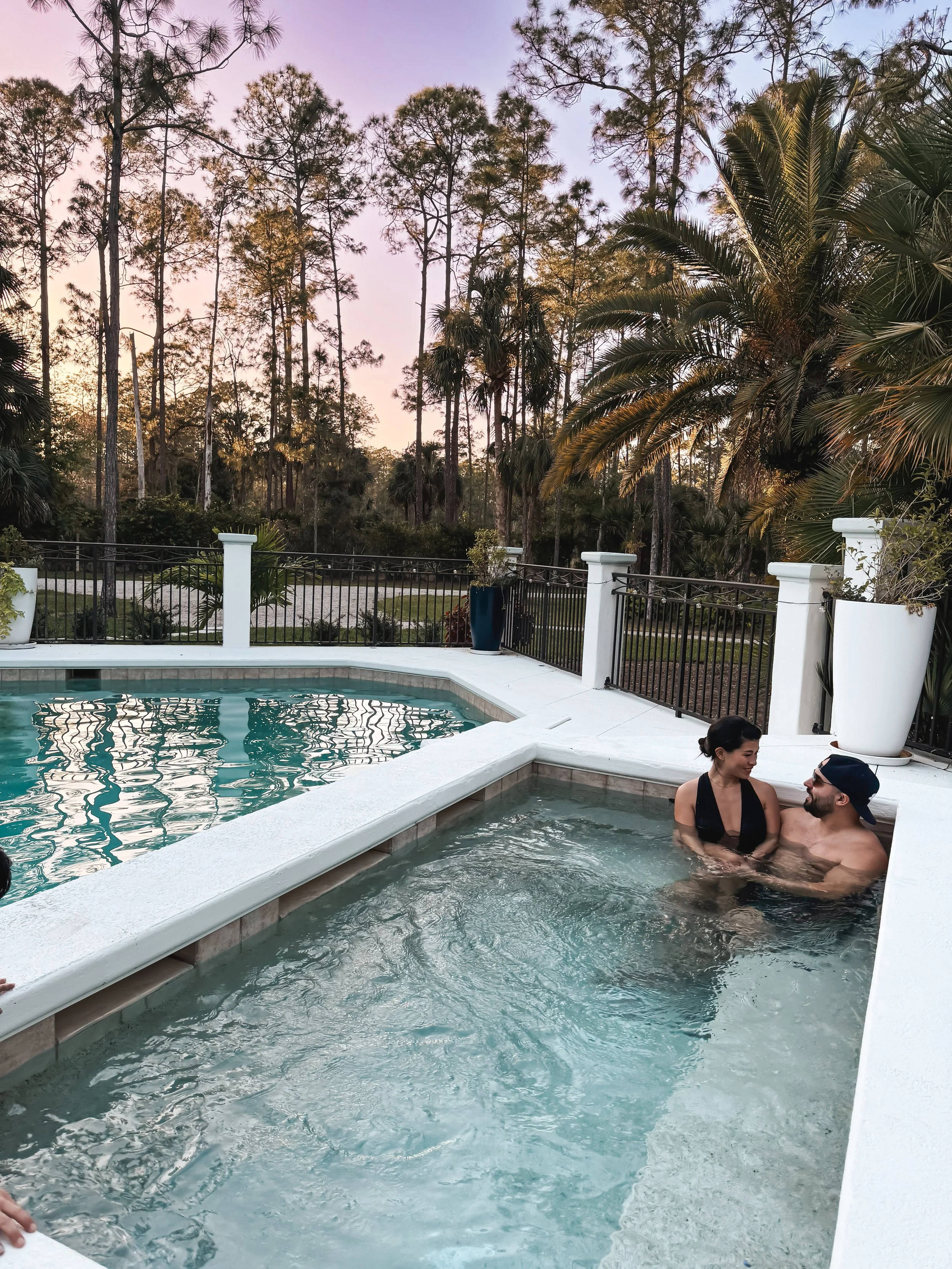 Two people are relaxing and chatting in a hot tub by a white patio fence, with tall trees and a purple sunset sky in the background.