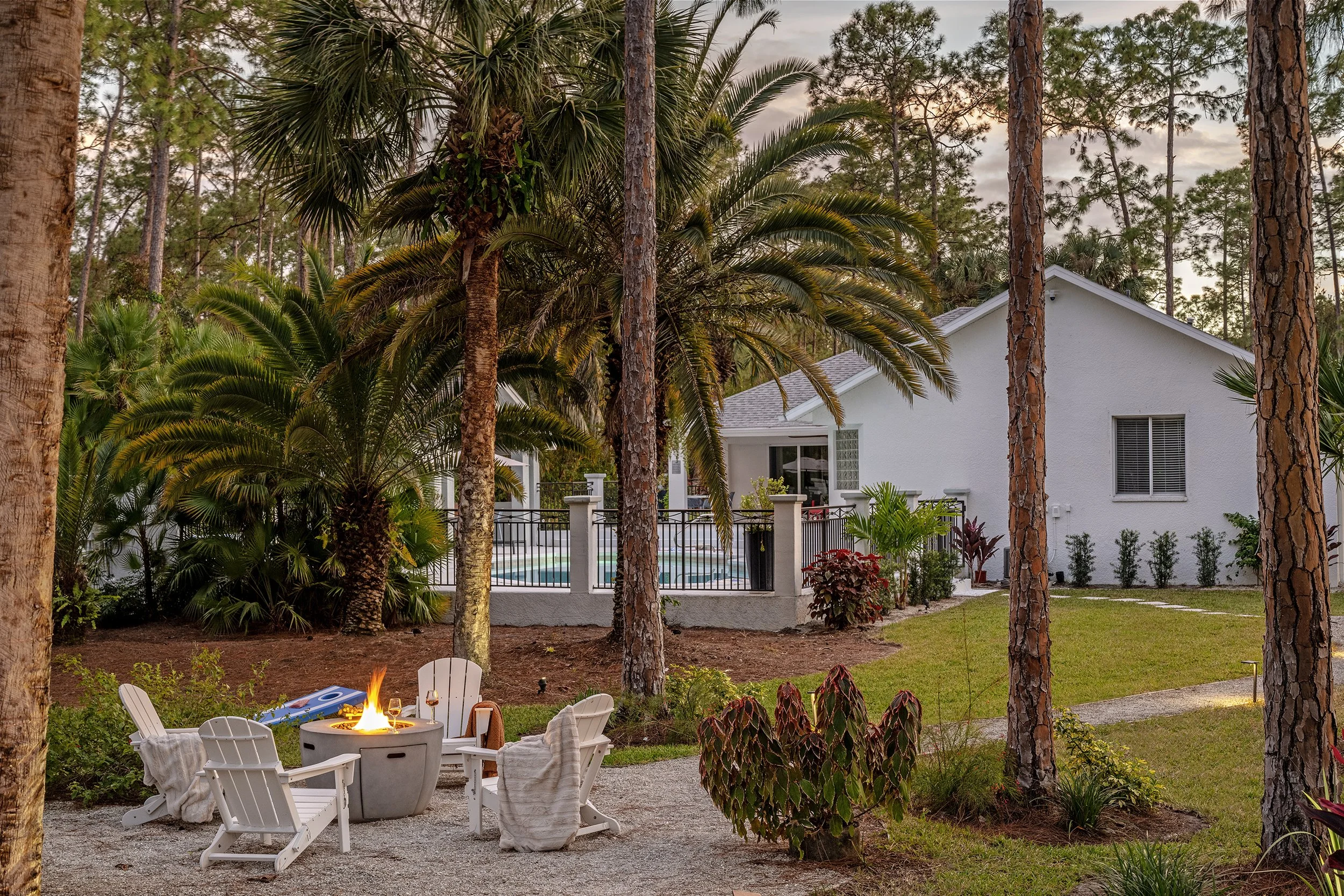 A backyard with a fire pit surrounded by white Adirondack chairs, green plants, and tall trees in front of a white house with a backyard pool.