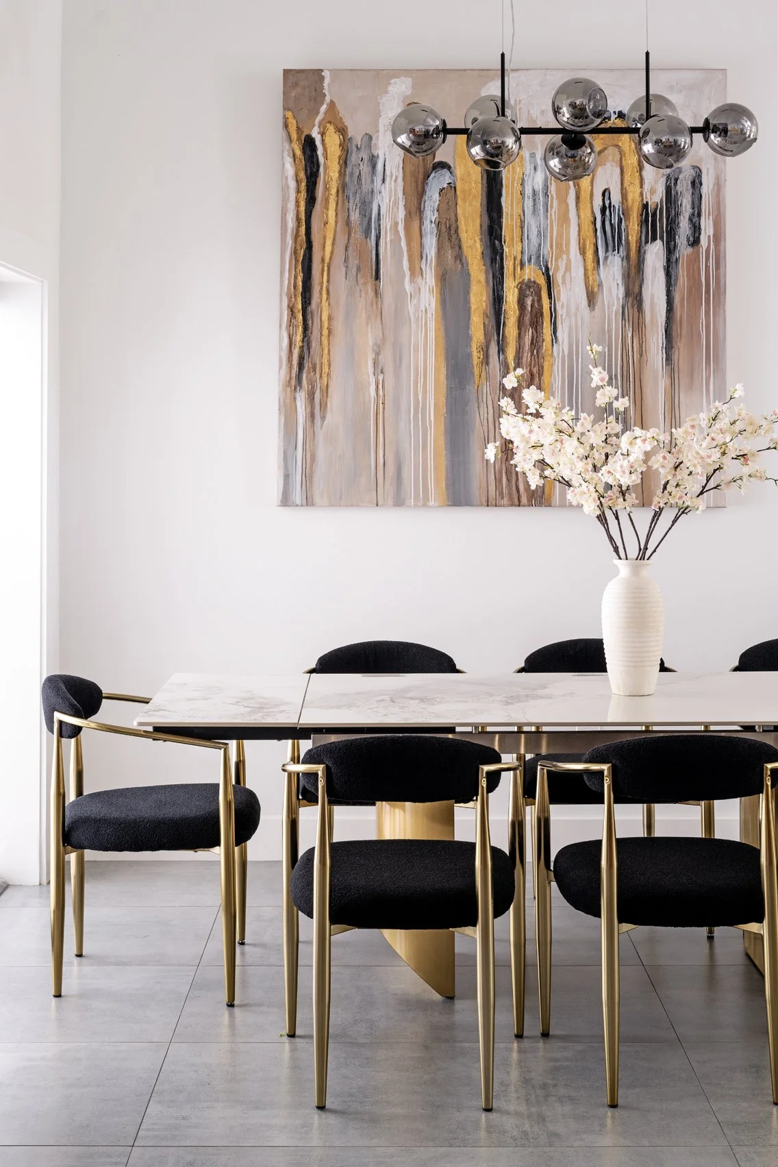 Modern dining room with a white marble table, black upholstered chairs with gold legs, a white vase with pink cherry blossoms, abstract wall art, and a black and chrome chandelier.