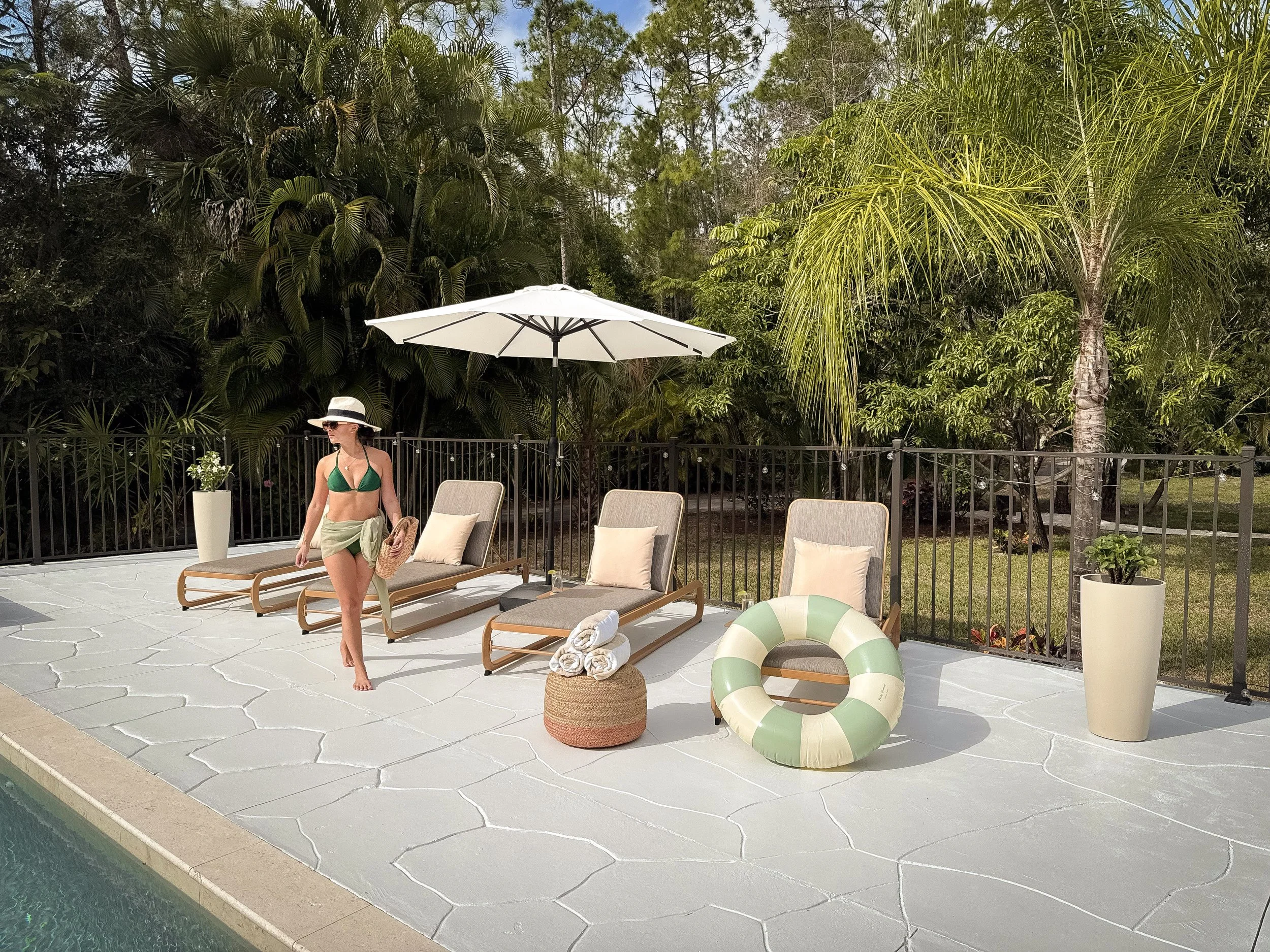 A backyard poolside area with pool in the foreground, a woman in a green bikini and hat walking, and lounge chairs with rolled towels, large white umbrella, potted plants, and tropical trees in the background.