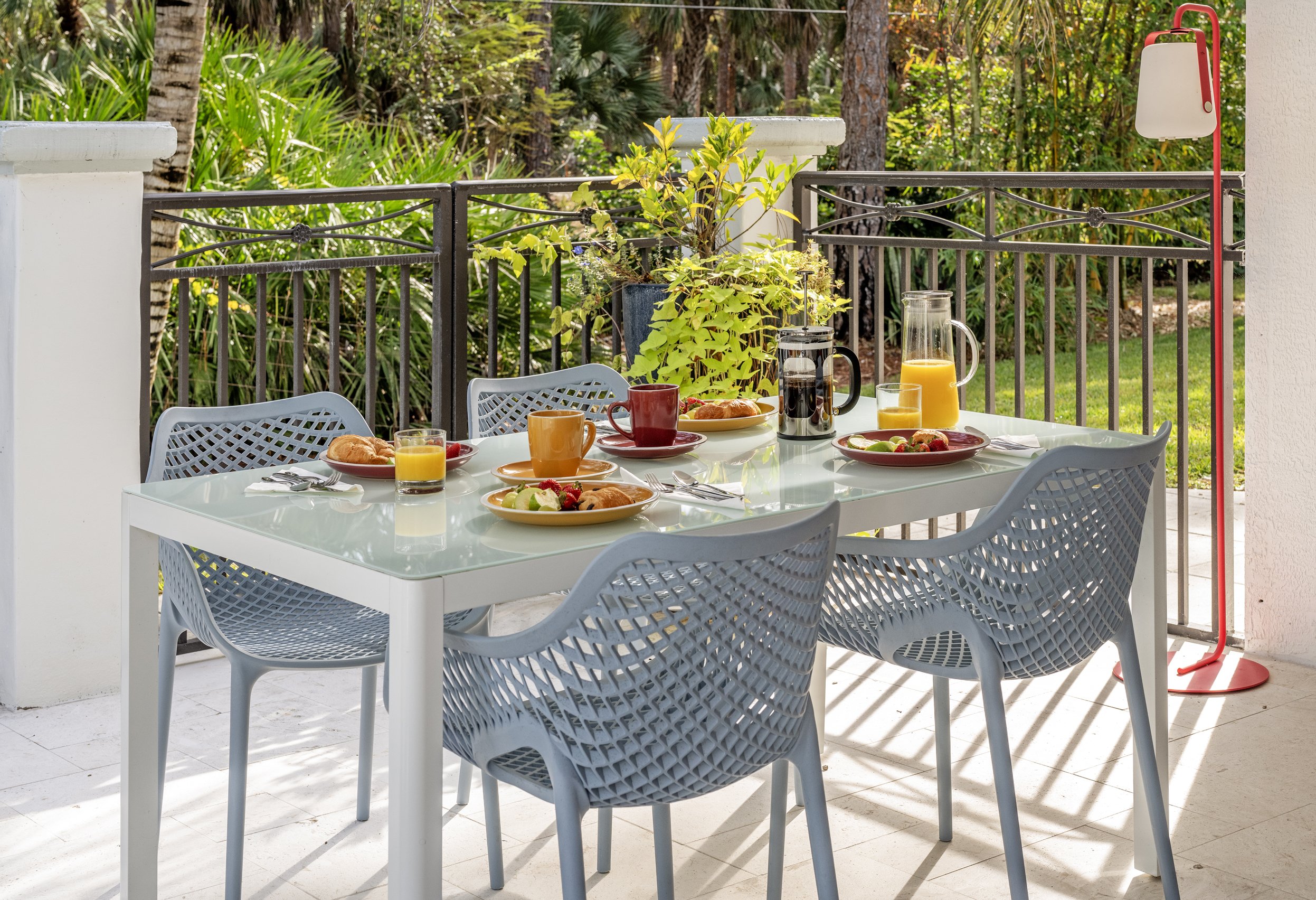 An outdoor dining table set for breakfast with plates of croissants, fruit, and glasses of orange juice. The table is surrounded by gray chairs, with a lush green forest in the background. There is a white and red lamp on the right side.