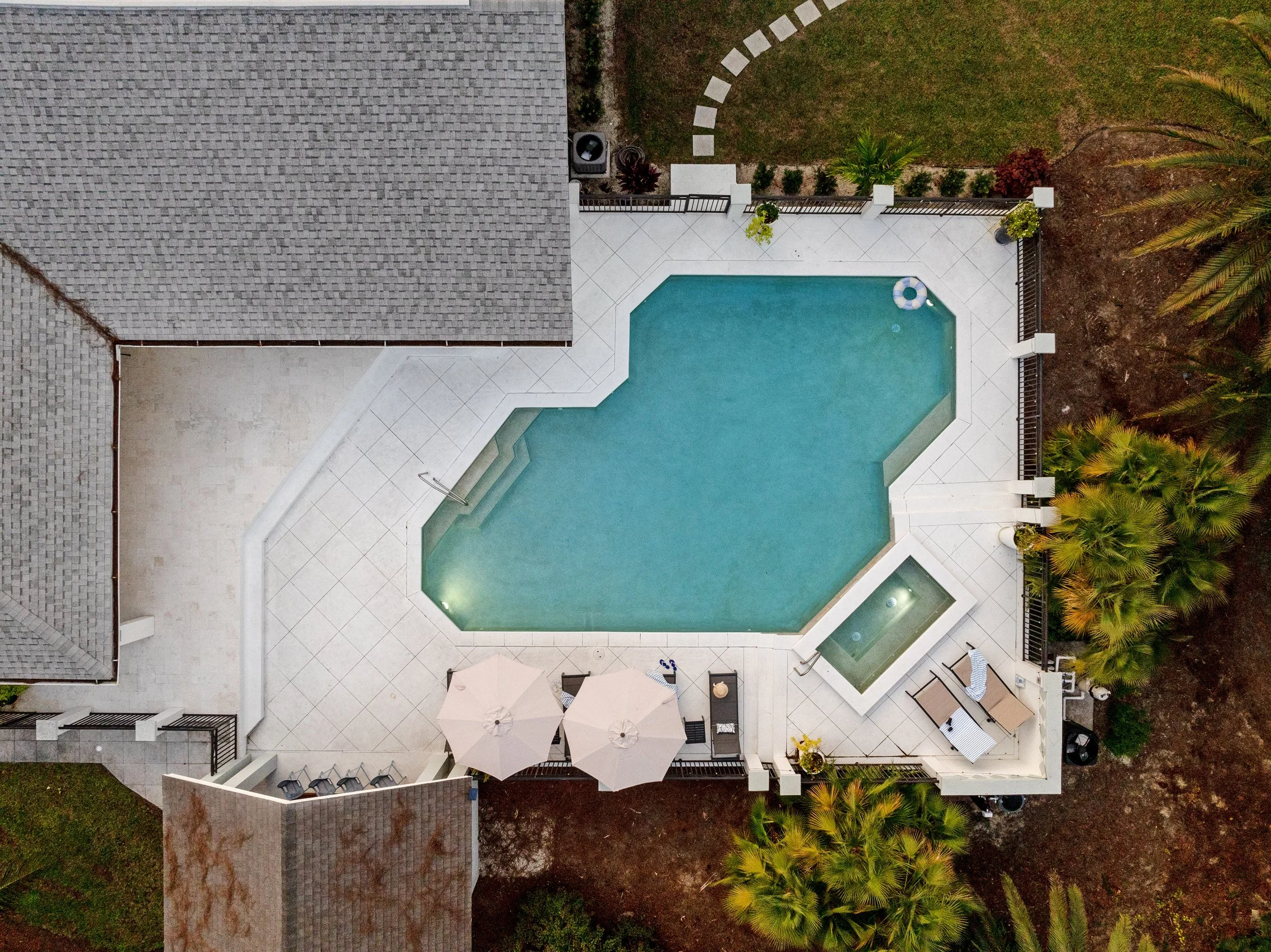 Aerial view of a backyard with a swimming pool, white tiled patio, outdoor table with umbrellas, lounge chairs, and surrounding greenery including palm trees.