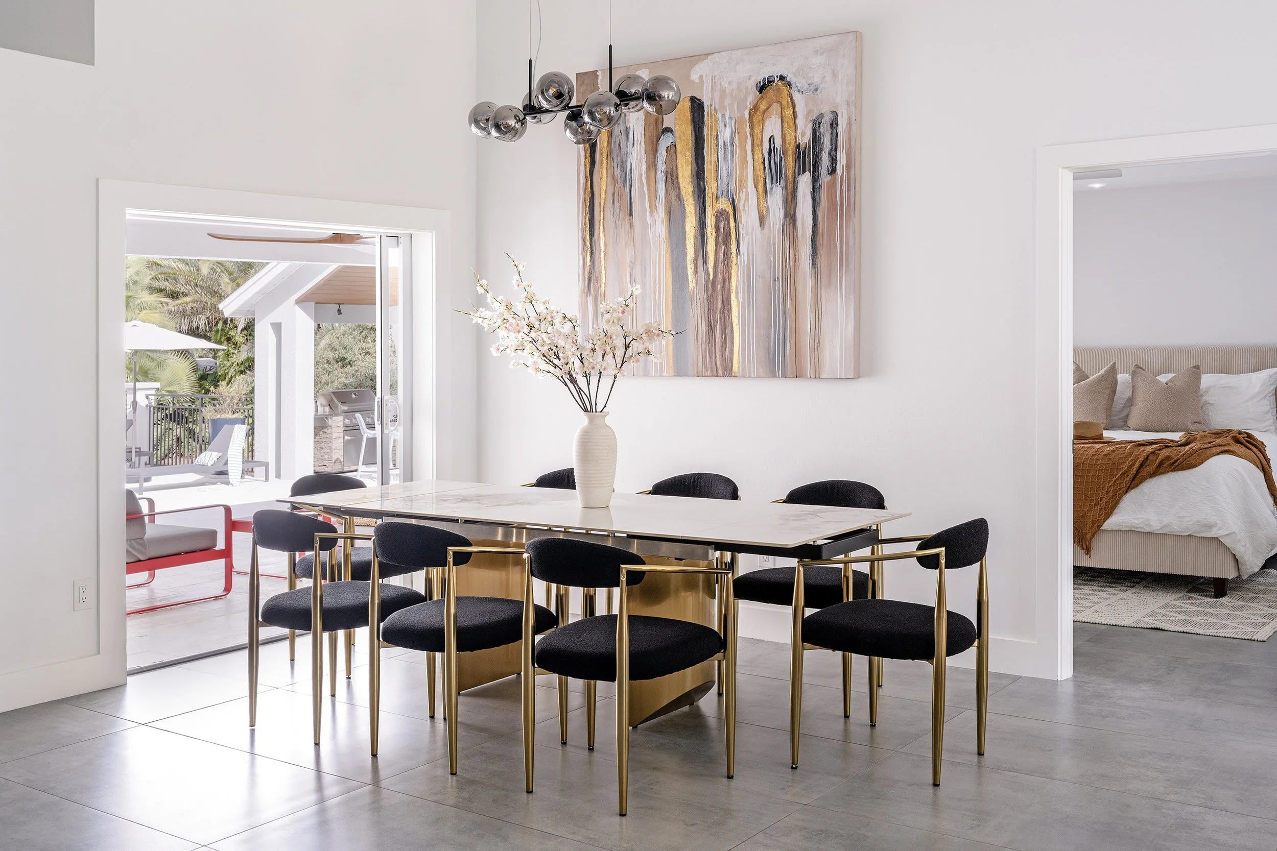 A modern dining area with a marble table surrounded by black chairs with gold legs, a white vase with pink cherry blossoms, a large abstract painting with gold, black, and white colors on the wall, and a contemporary black chandelier with multiple glass globes hanging from the ceiling. To the right, a bedroom with a bed with a beige headboard, white bedding, and beige pillows is visible through a doorway. The space is bright with natural light coming through an open sliding door leading to a patio with greenery and outdoor furniture.