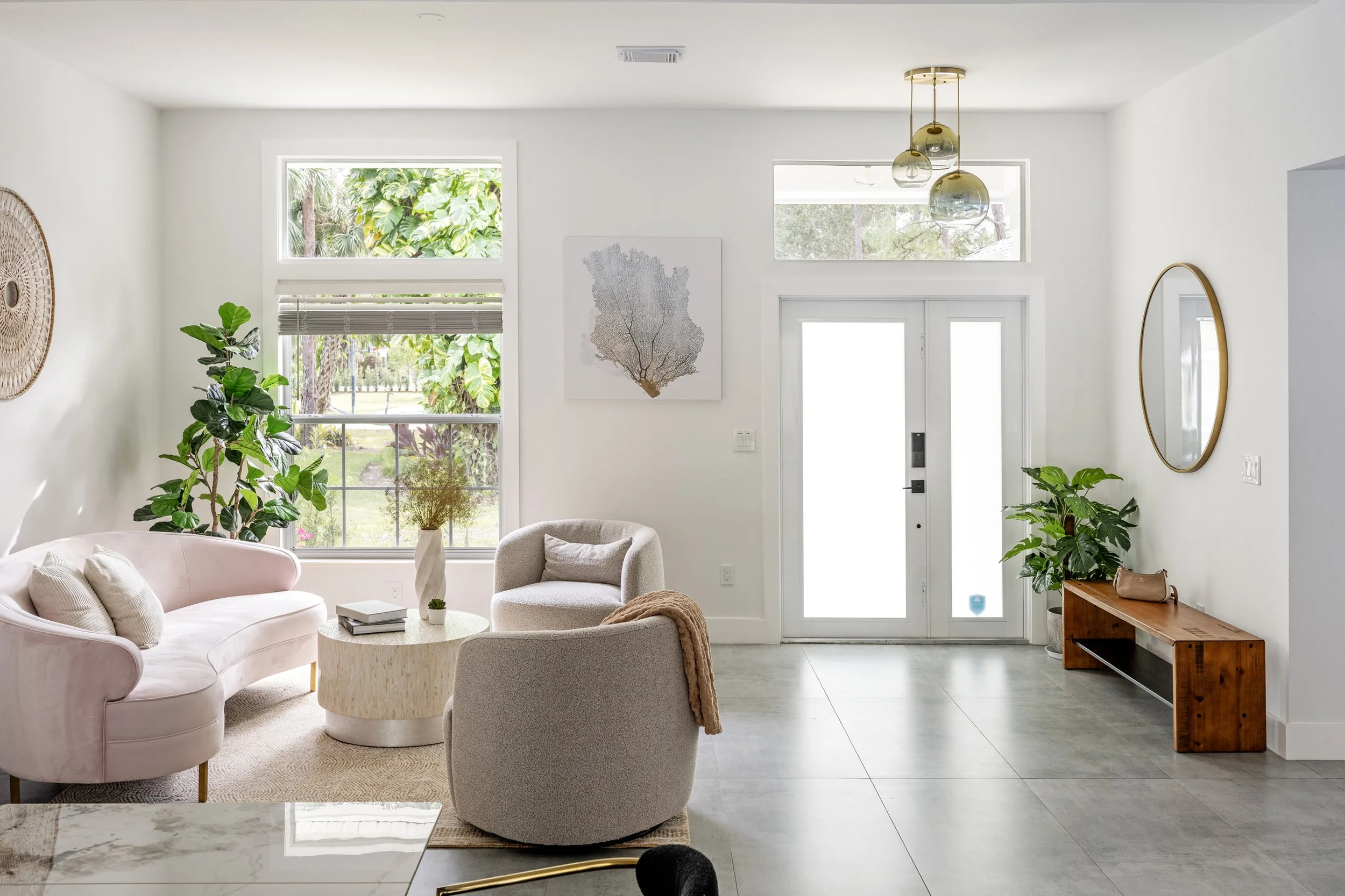 A bright living room with white walls and large windows showing trees outside. The room has a pink sofa, a beige armchair, a circular coffee table, and various plants including a tall fiddle leaf fig. Decor includes a wall art piece resembling coral 