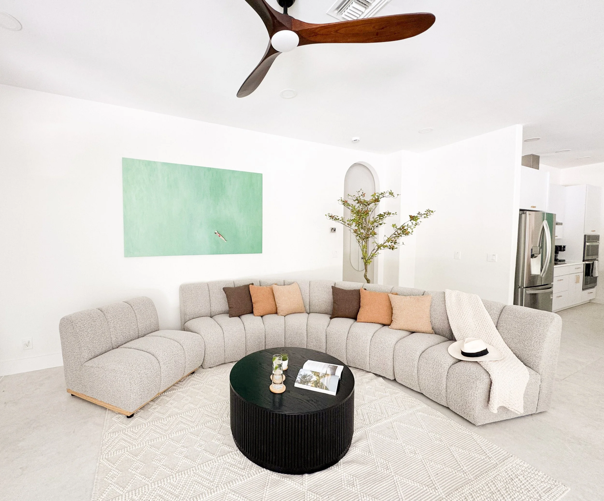 Living room with a large L-shaped beige sofa with brown and beige pillows, black round coffee table with drinks and magazines, beige rug, white walls with green painting, ceiling fan, potted plant, and kitchen appliances in background.