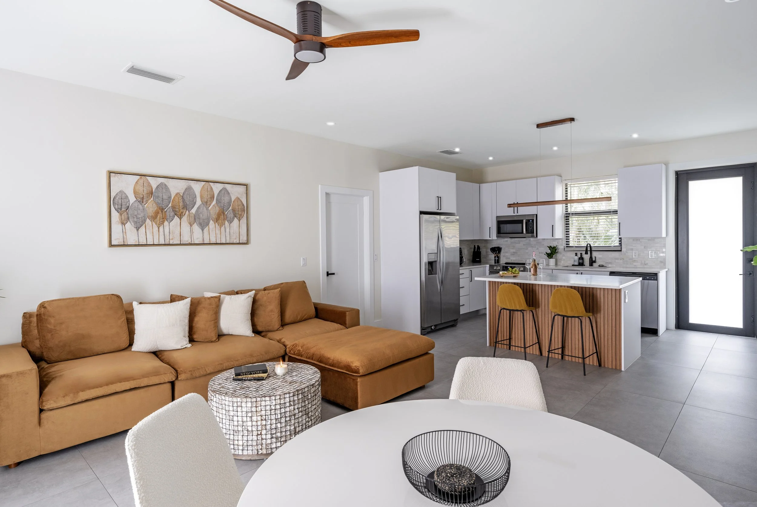 Open-concept living and kitchen area with a brown sofa, white walls, a round coffee table, and a modern kitchen with white cabinets, stainless steel refrigerator, and bar seating with two yellow chairs. Ceiling fan and decorative wall art are also vi