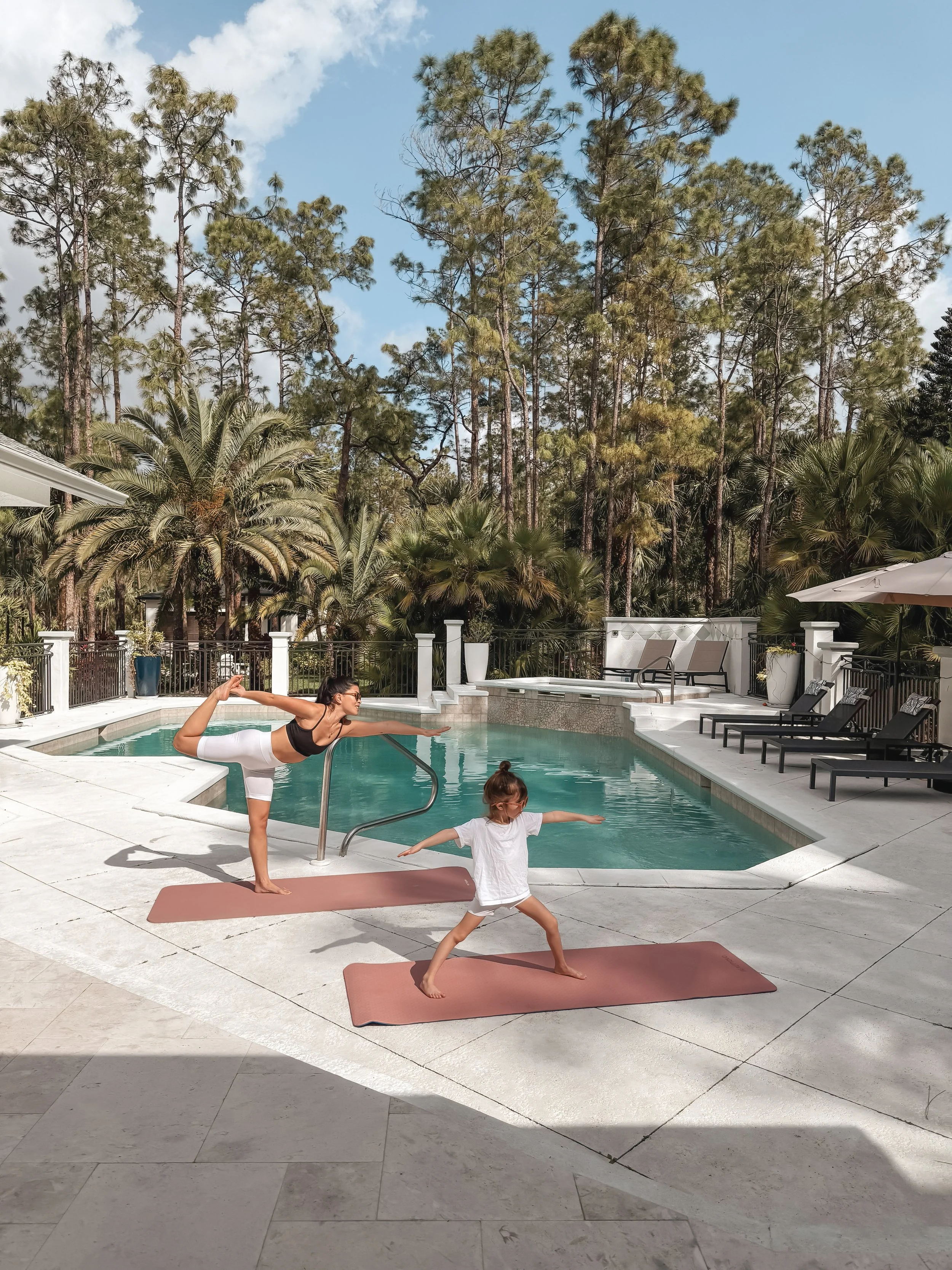 A woman and a young girl are practicing yoga on mats beside a swimming pool. The woman is holding a yoga pose with one leg raised and extended behind her, while the girl is in a warrior pose. The pool area is surrounded by lounge chairs, palm trees, 
