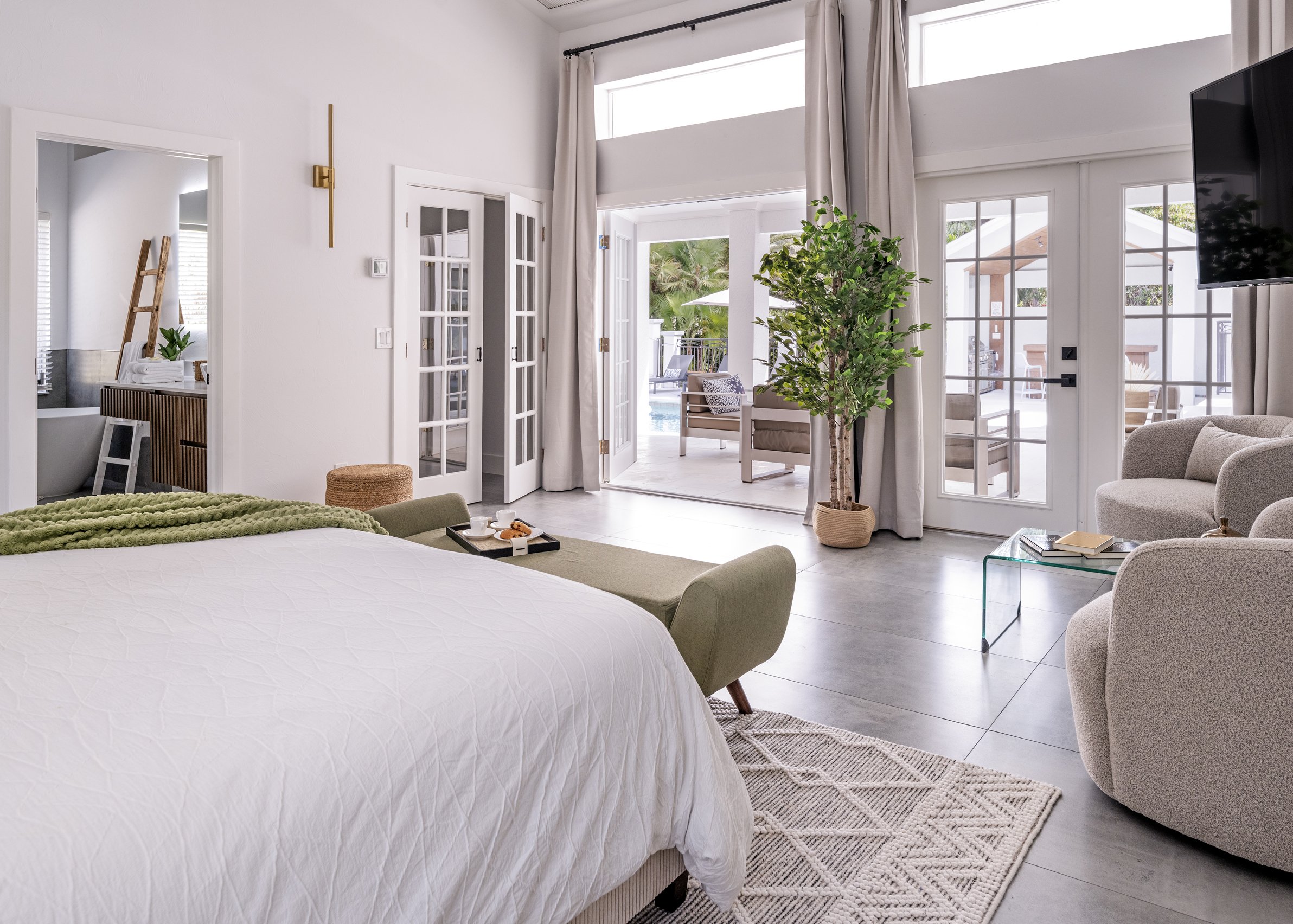 Bright living room with white walls, French doors leading outside to a patio with seating and a pool, large window, green potted plant, and modern furniture.