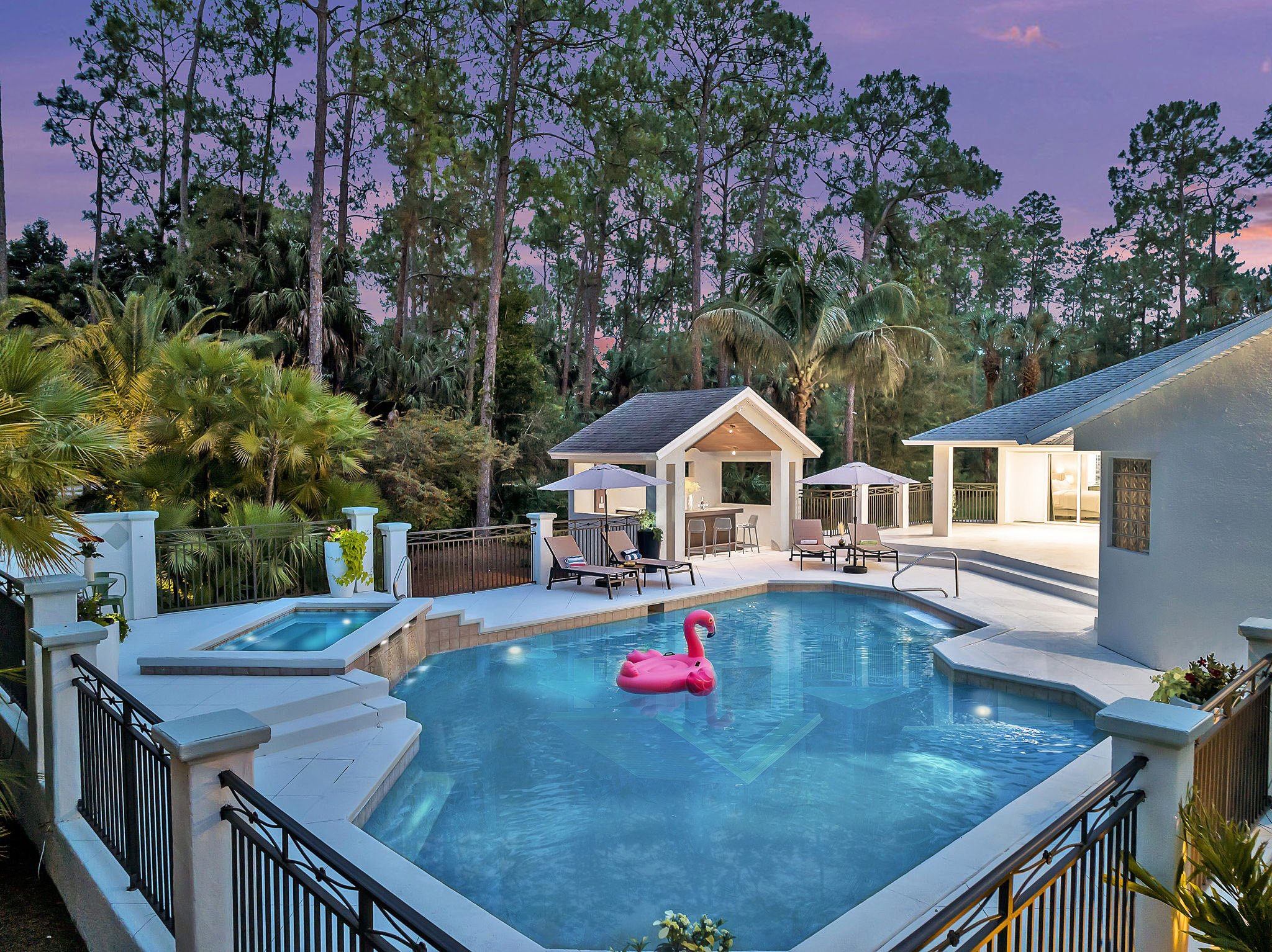 A backyard swimming pool with a pink flamingo float and surrounding lounge chairs, enclosed by a black metal fence, with a backdrop of tall trees and a purplish evening sky.