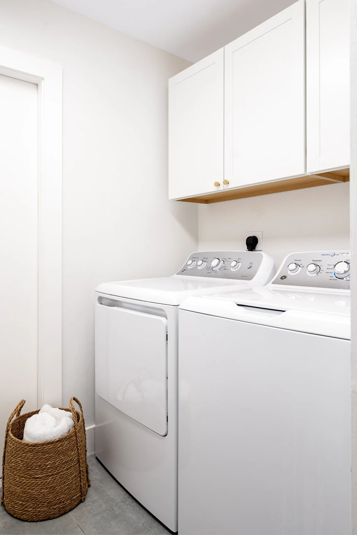 A laundry room with a white washer and dryer side by side, white cabinets above, a basket with towels on the floor, and a wall outlet with a plug.