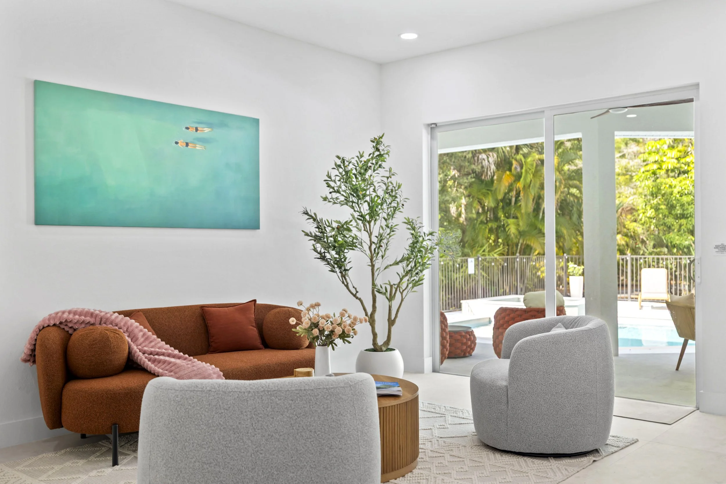 Living room with a rust sofa, pink throw blanket, glass door leading to an outdoor patio with outdoor furniture, a large potted plant, and abstract wall art.