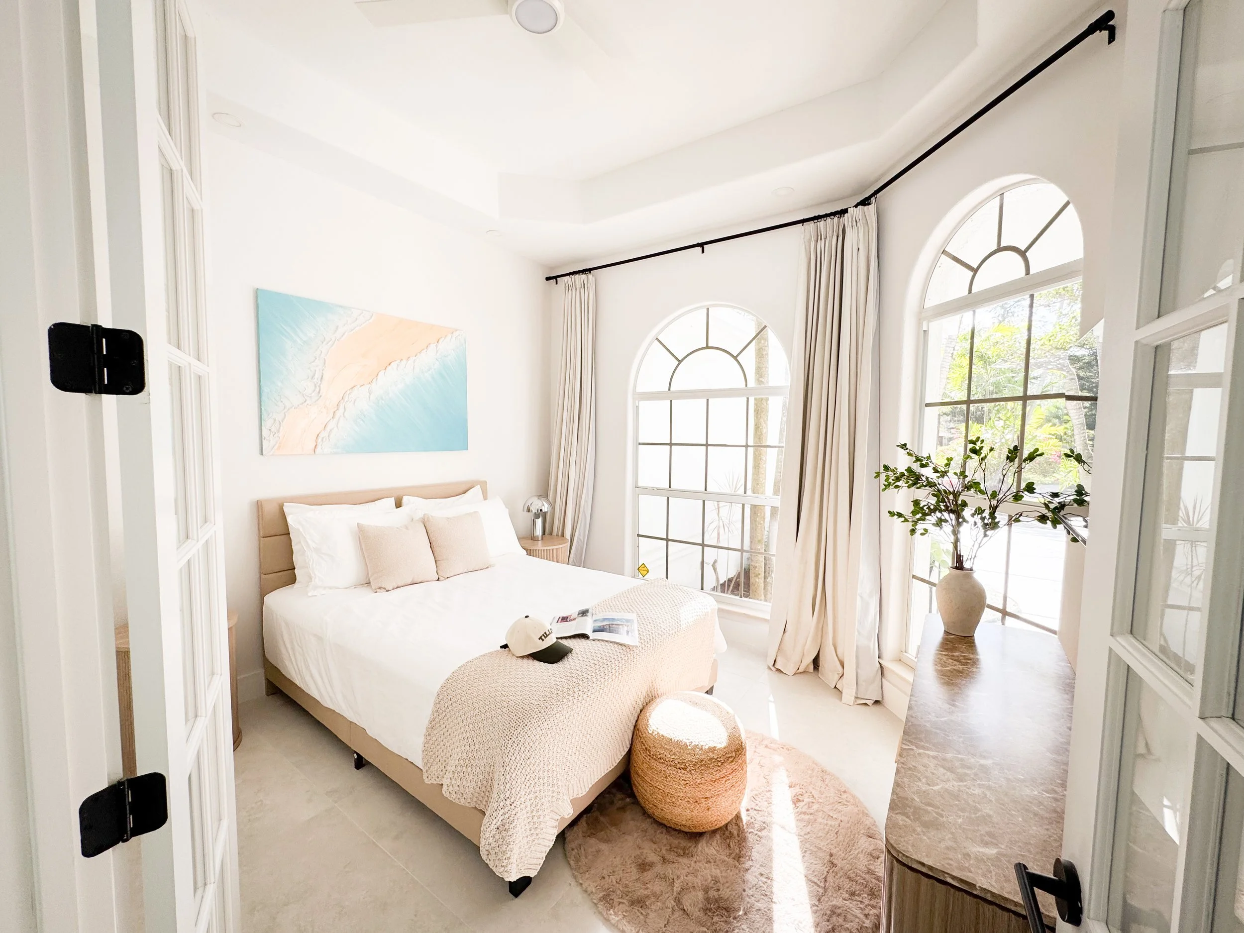 Bright bedroom with white walls, large arched windows with curtains, a white bed with beige pillows, a painting on the wall, and a potted plant near the window.