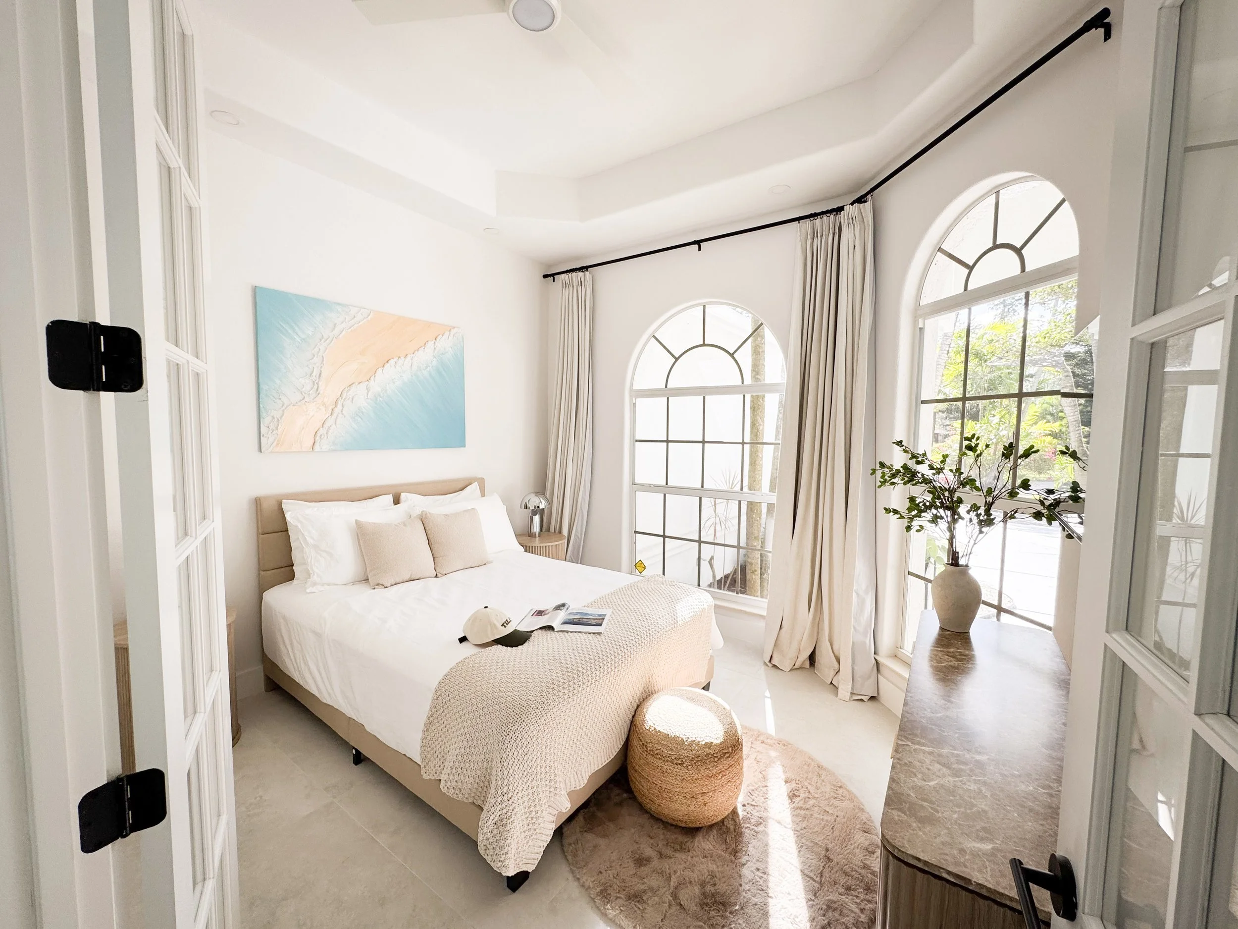 A bedroom with large arched windows, white walls, a bed with beige headboard, white bedding, and decorative pillows, a small round ottoman, and a potted plant near the window.