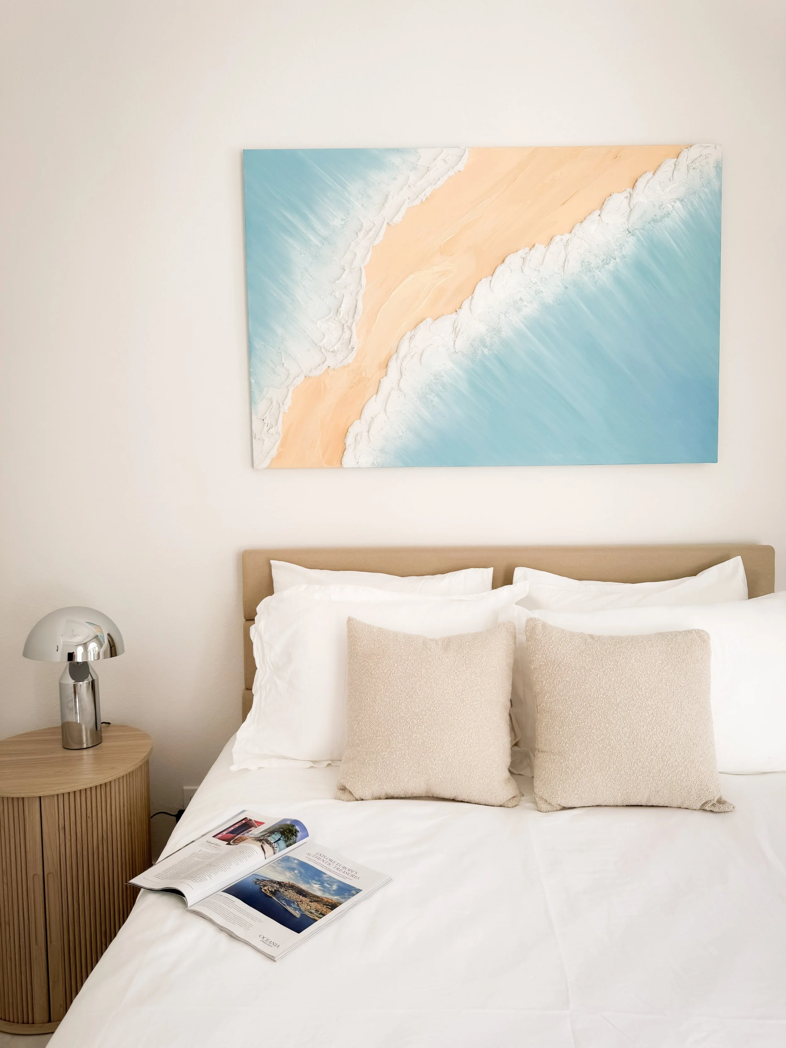 A neatly made bed with beige pillows, a wooden nightstand with a silver lamp, a magazine open on the white bedsheet, a colorful abstract painting above the bed.