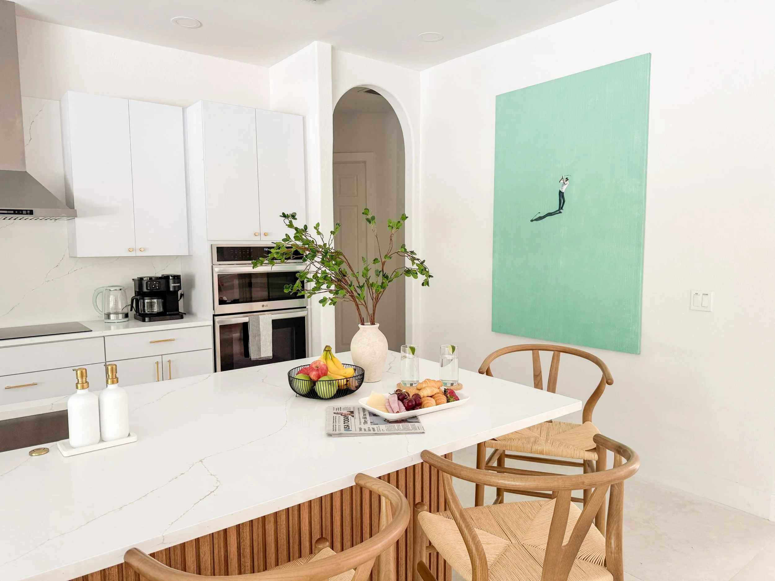 Modern kitchen with white cabinets, a built-in oven, and a coffee maker. A white island countertop has a fruit bowl, a newspaper, and soap dispensers, with dining chairs nearby. A large green painting hangs on the wall, and there's a potted plant on 