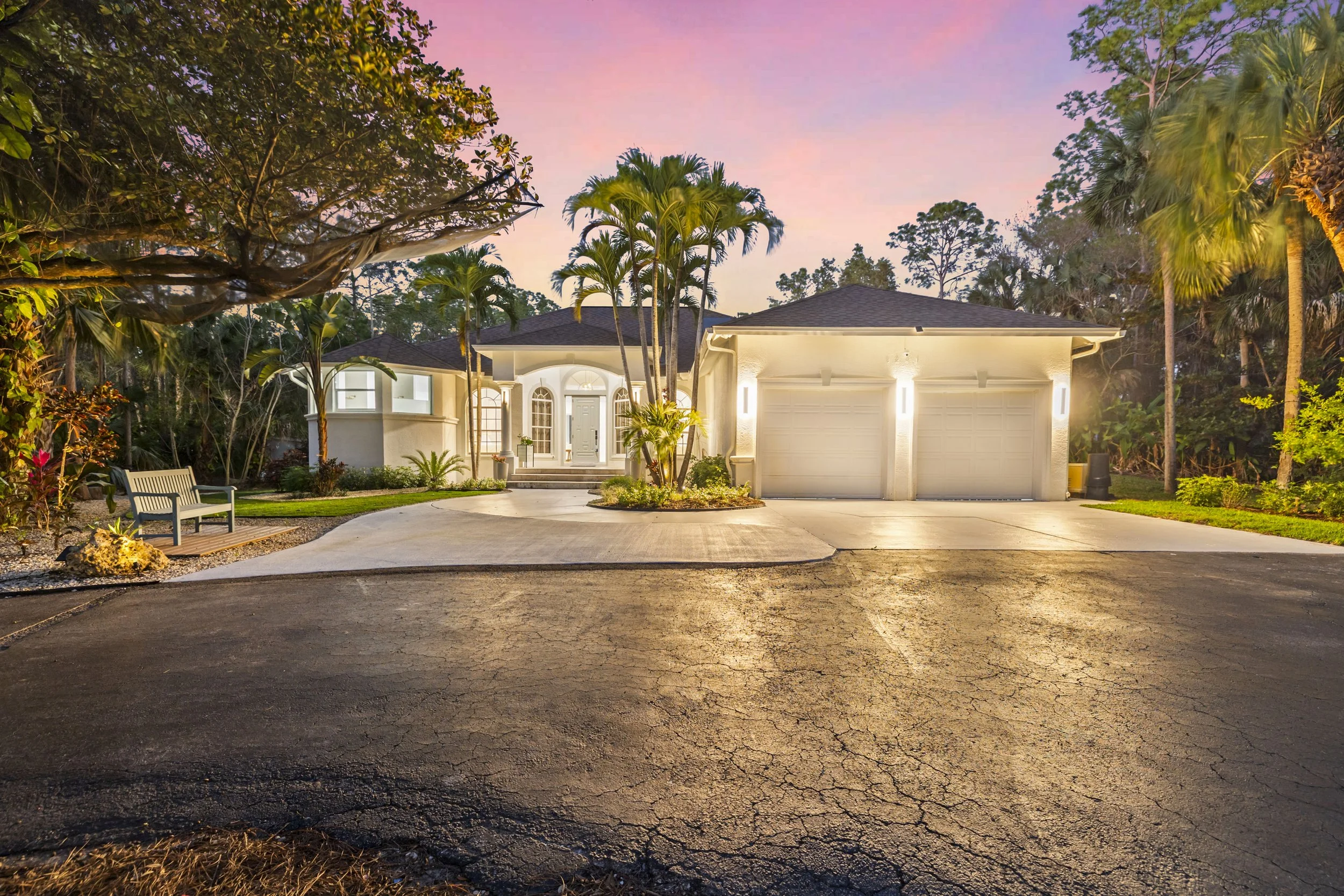 A well-lit house with a driveway and surrounded by trees and plants during sunset or twilight.