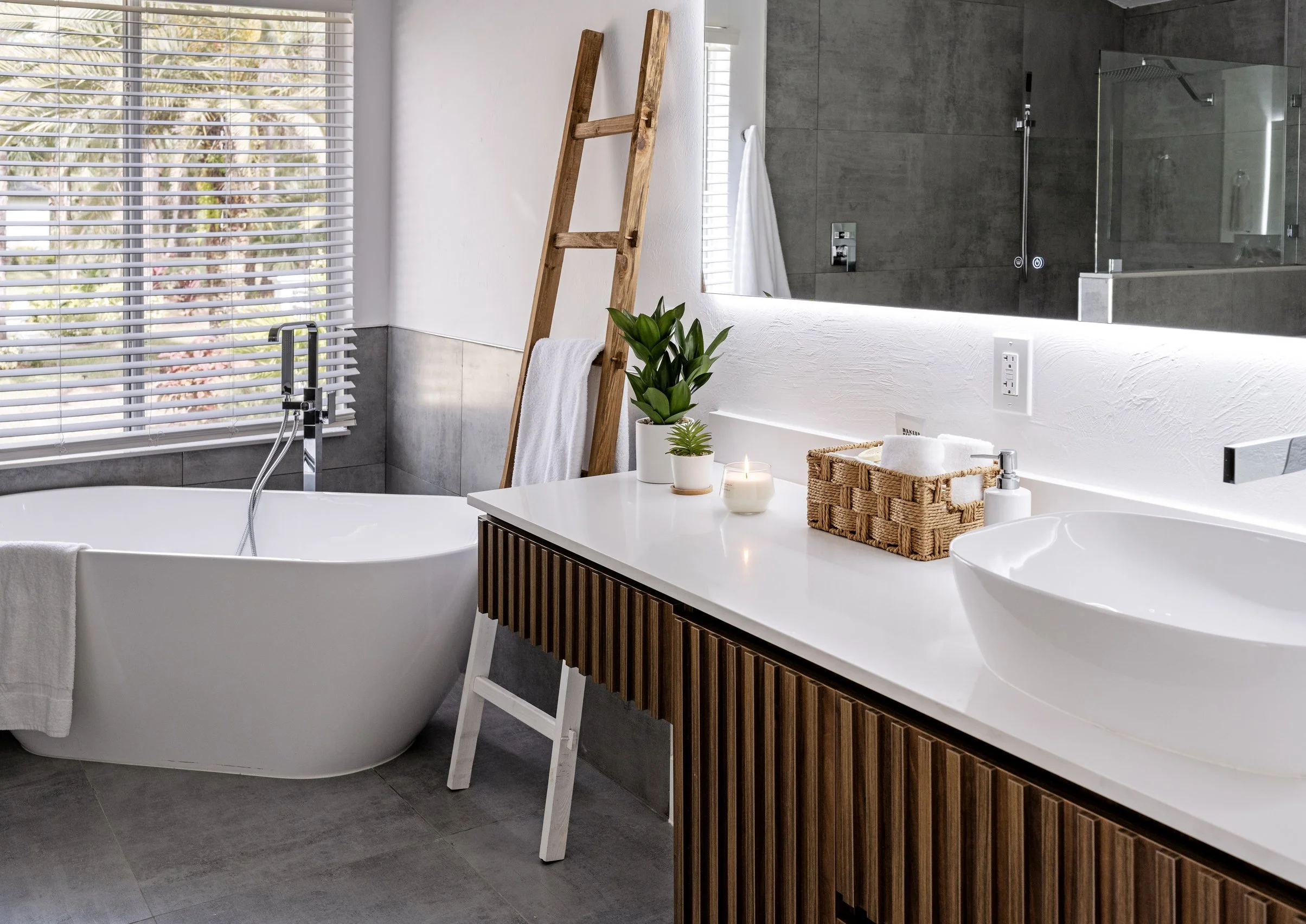 Modern bathroom with a freestanding white bathtub, large mirror, wooden ladder with a towel, green plants, candles, and a vanity with a sink and soap dispenser.