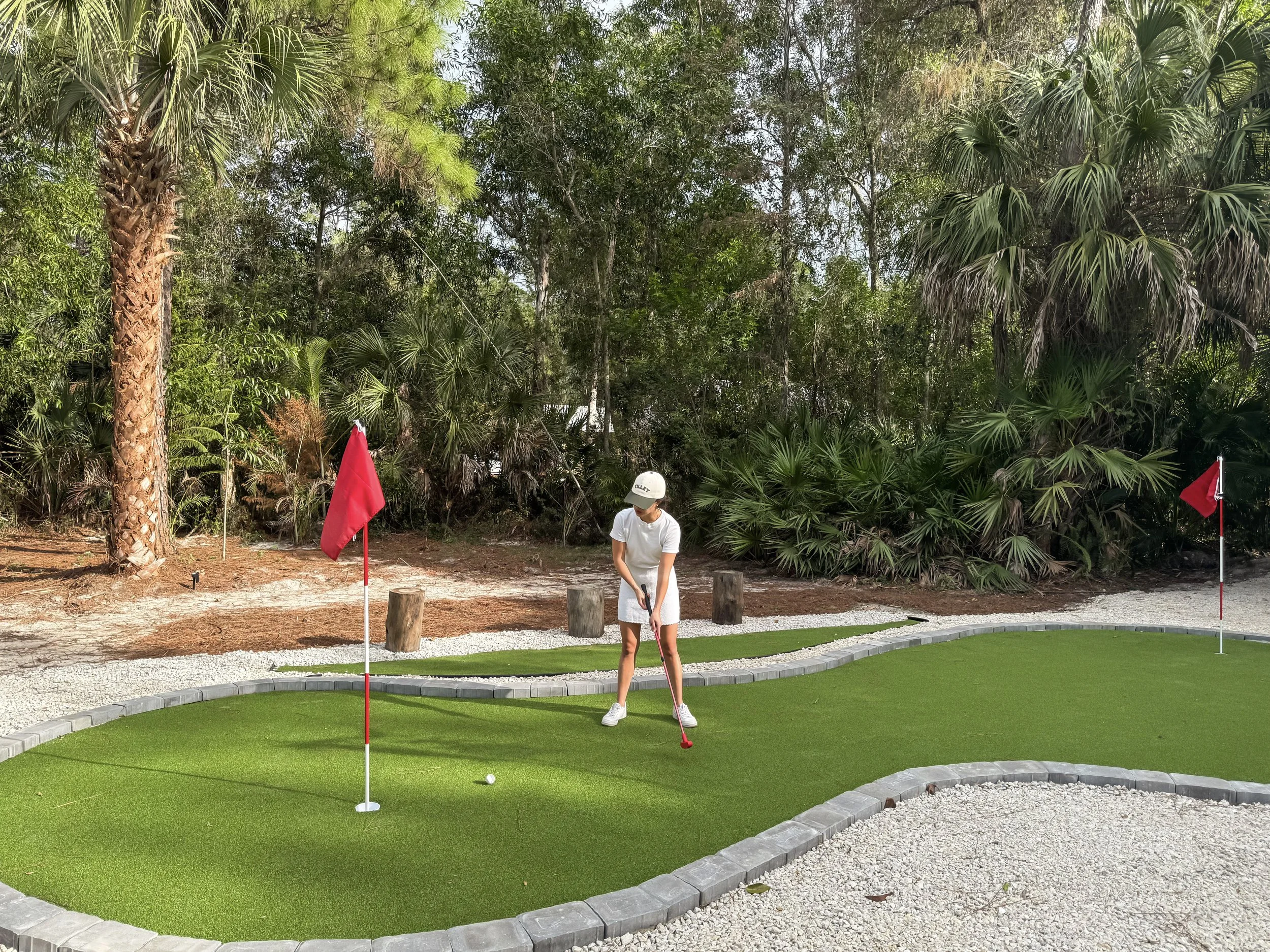 A woman playing mini golf on a course with green artificial turf, surrounded by a stone border. She is wearing white clothing and a white cap, preparing to putt the golf ball. In the background, there are tall trees and lush greenery, with red flags 