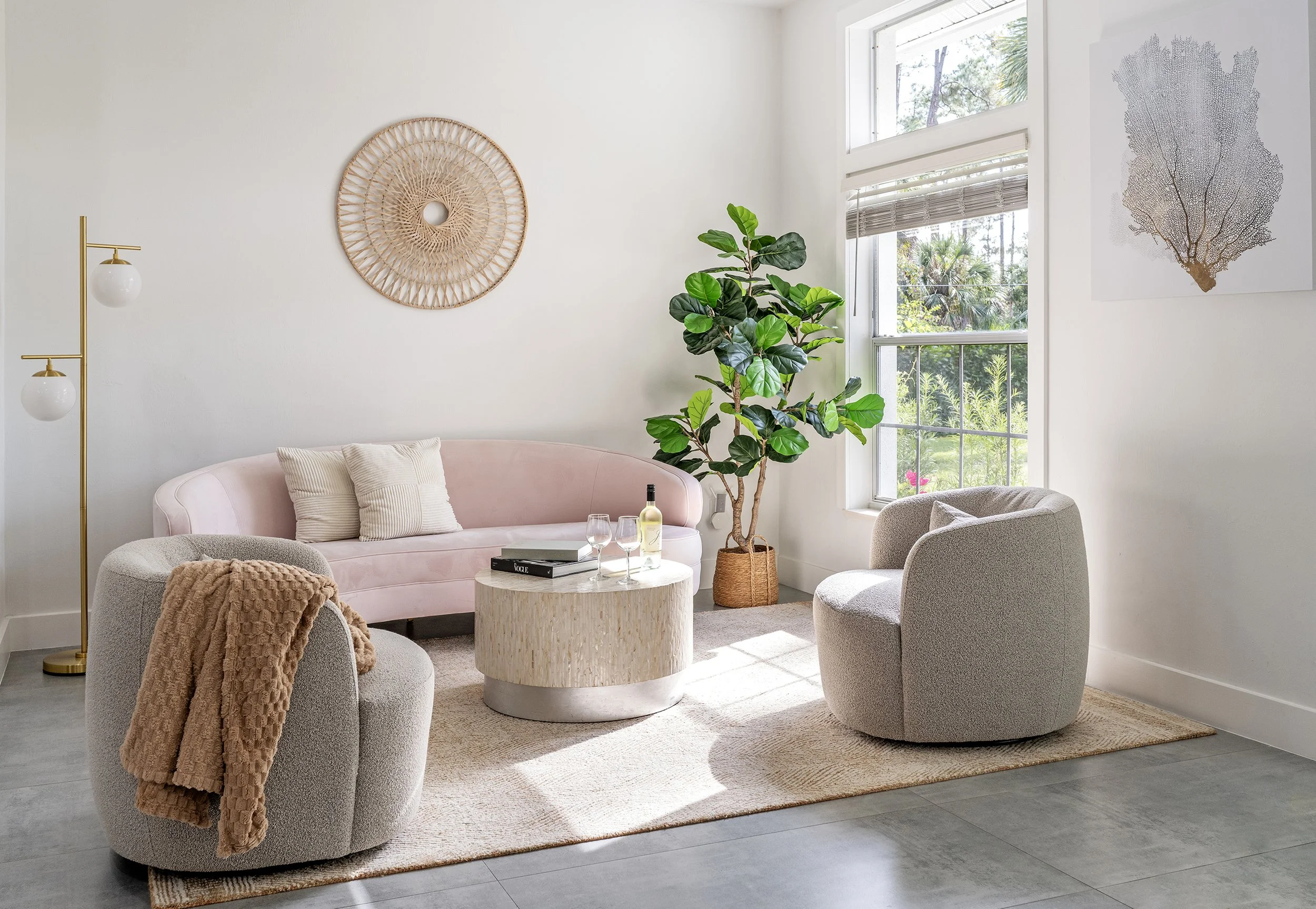 Bright living room with a pale pink sofa, gray armchair, white textured coffee table, and large green houseplant near a window with white blinds.